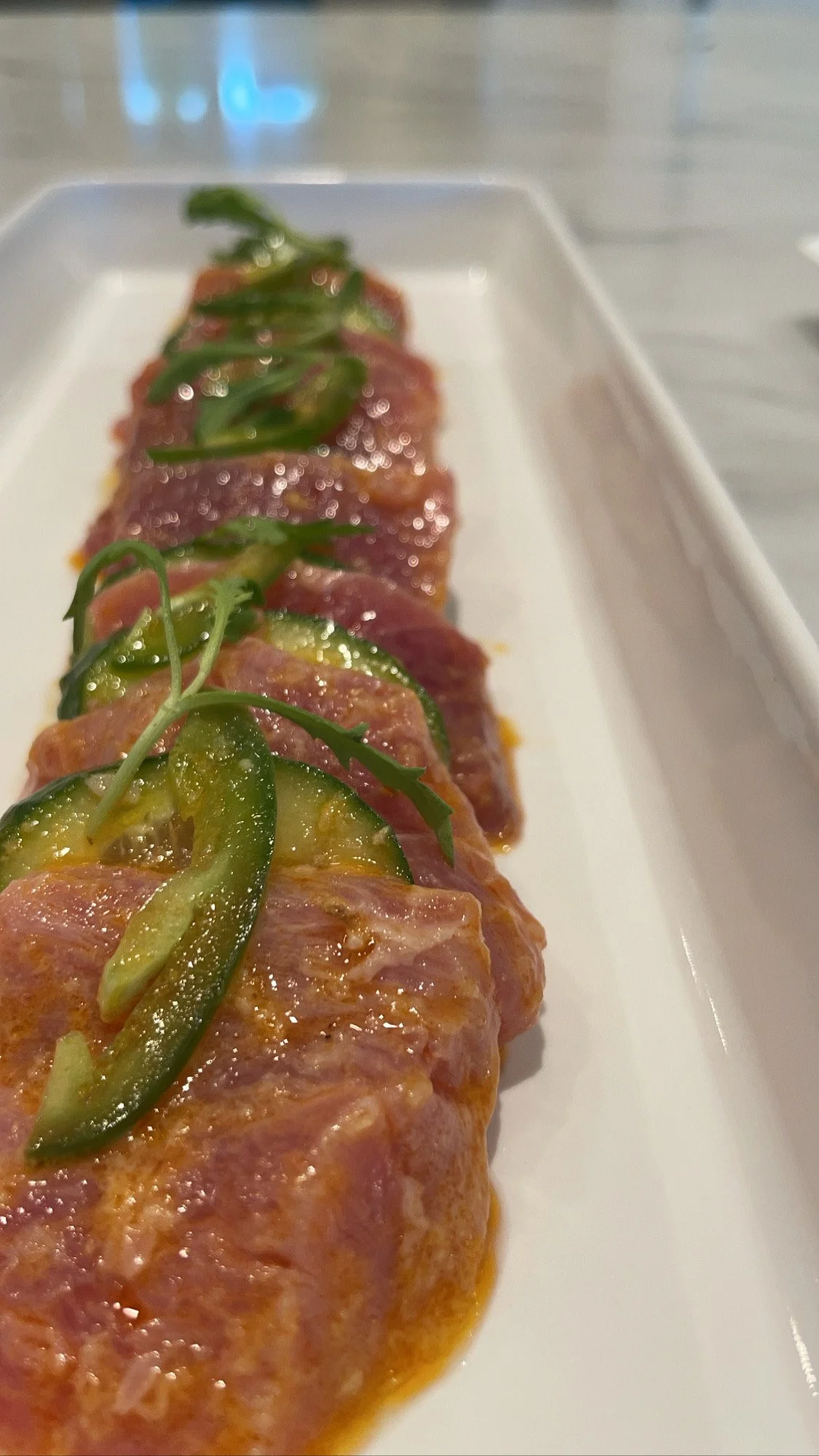 Close-up of sliced raw tuna topped with cucumber slices and microgreens, served on a white rectangular plate.