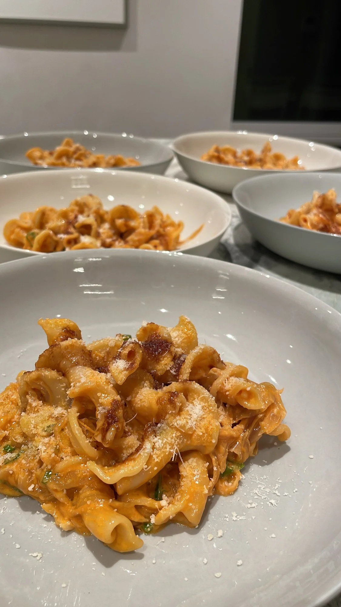 Fettuccine pasta with tomato sauce and grated cheese on a white plate, with multiple bowls of similar pasta in the background.