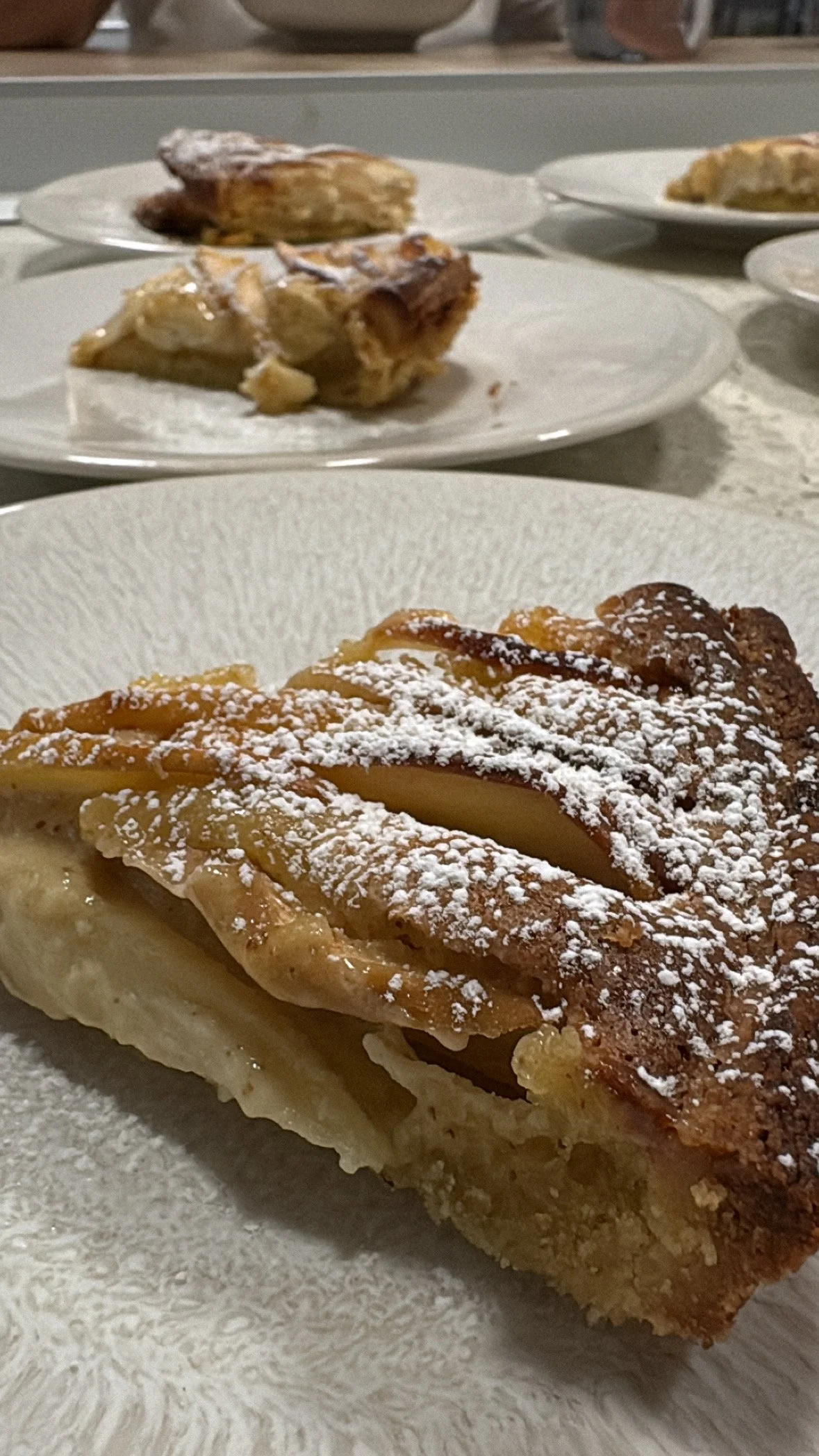A close-up of a slice of apple pie topped with powdered sugar, with other pies in the background on white plates.
