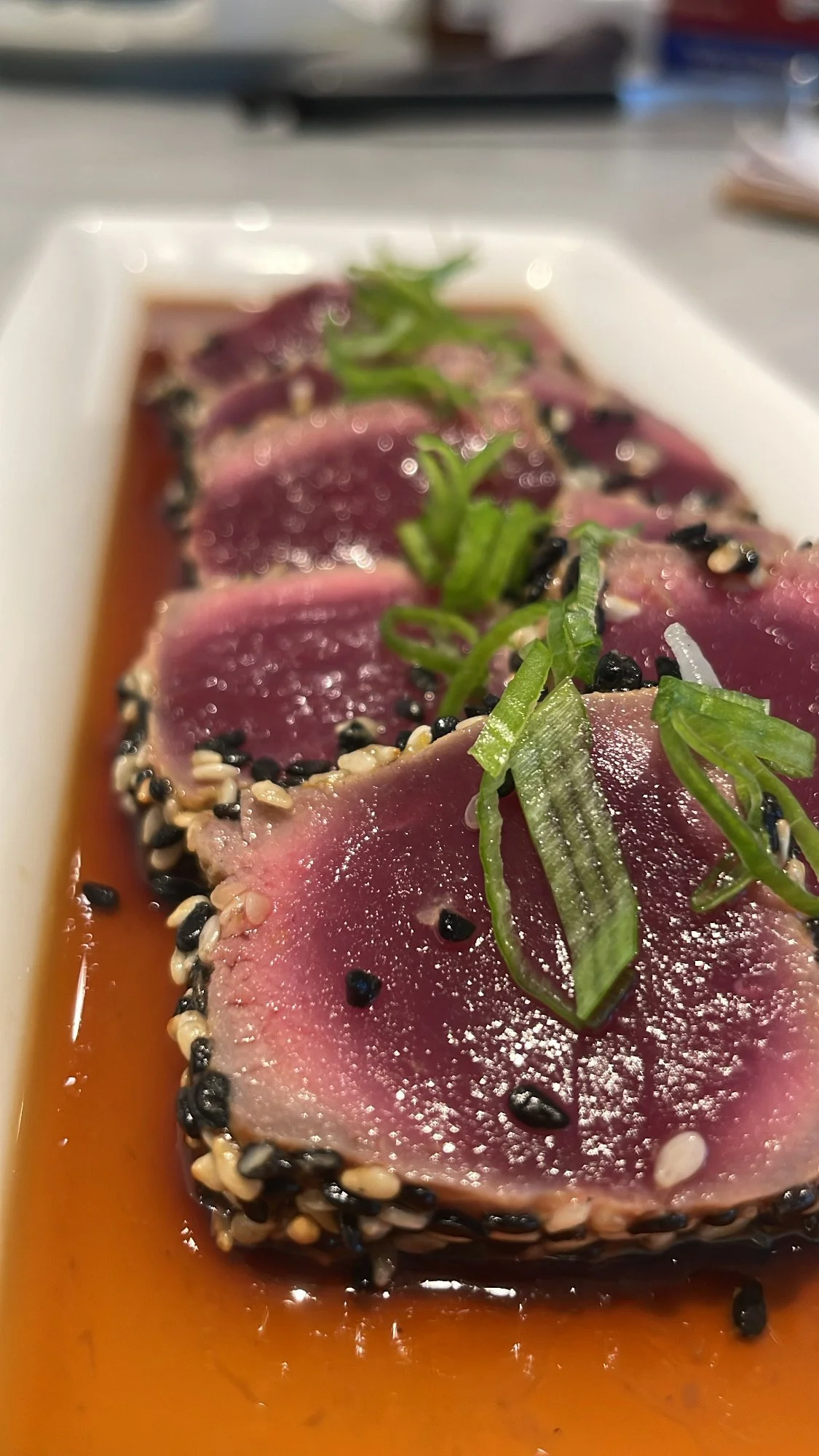 Close-up of ahi tuna sashimi topped with sliced green onions, sesame seeds, and black pepper, served with soy sauce.