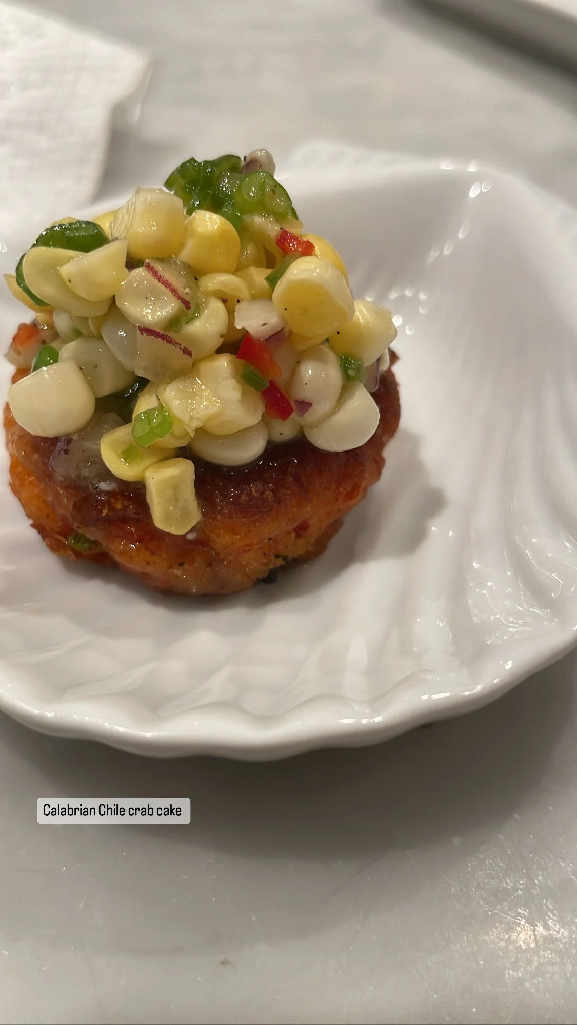 Calabrian Chile crab cake topped with a corn, red bell pepper, and green onion relish on a white plate.