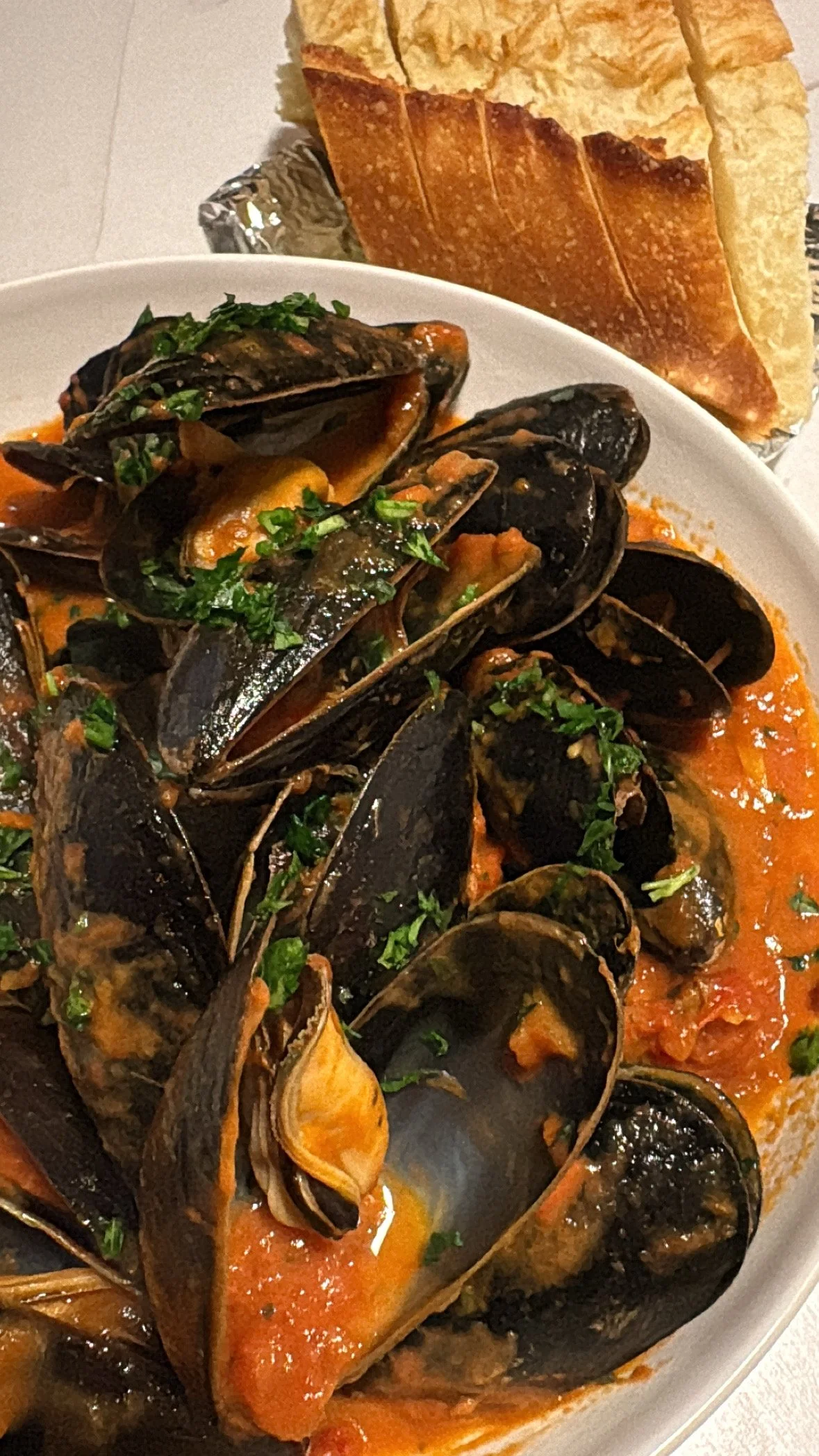 A white bowl of cooked mussels in tomato sauce garnished with chopped parsley, accompanied by slices of French bread.