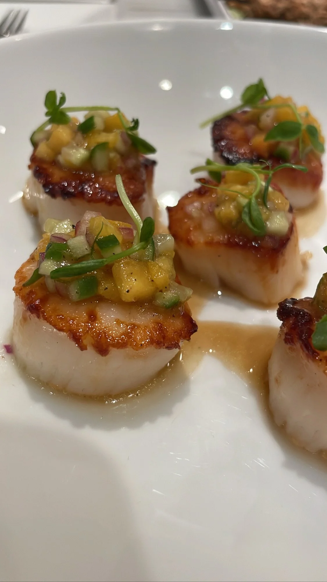 Close-up of seared scallops topped with diced green onions, pineapple, and microgreens on a white plate.