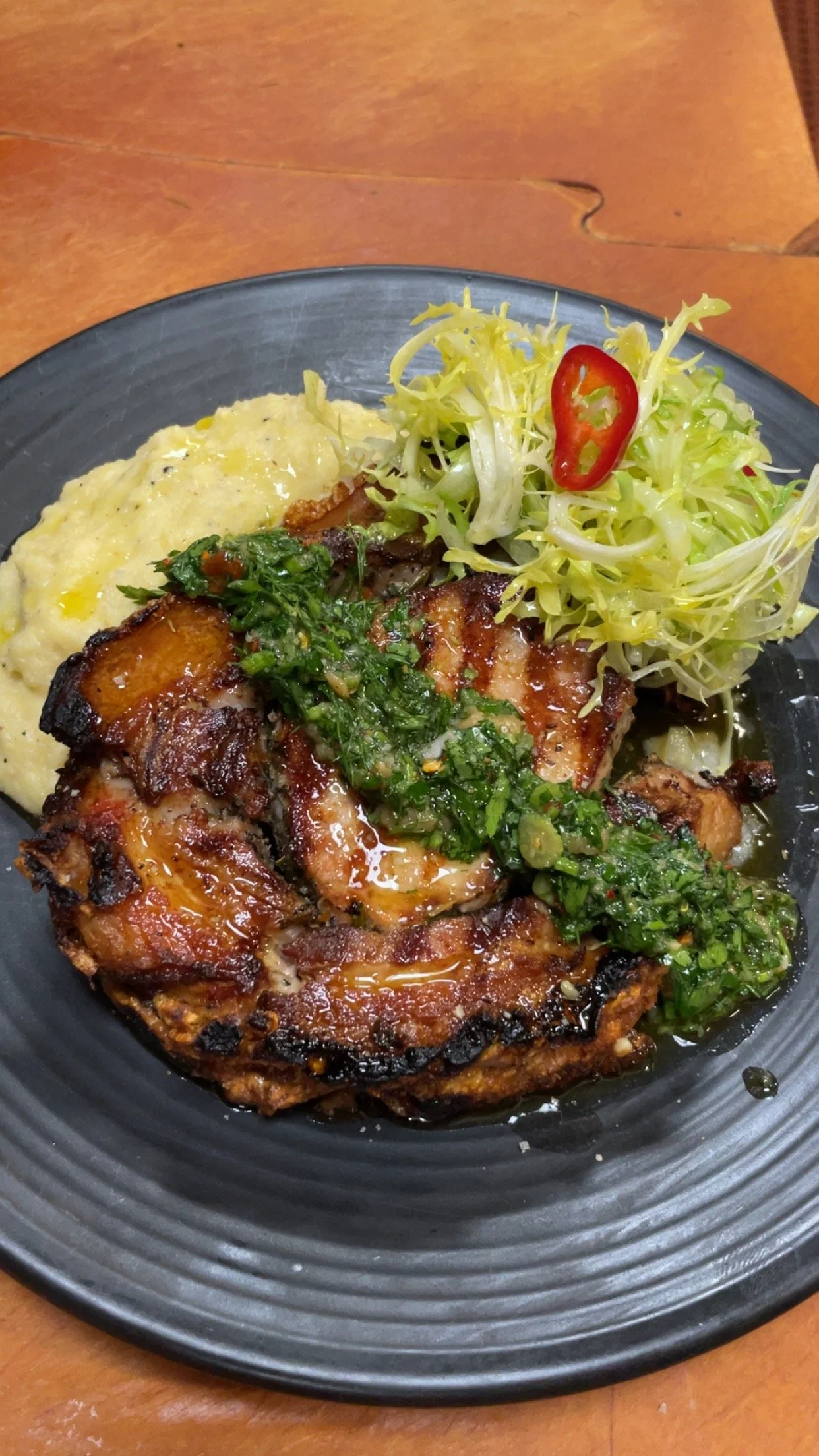 Grilled meat with green herb sauce, mashed potatoes, and side salad with lettuce and sliced chili pepper on black plate.
