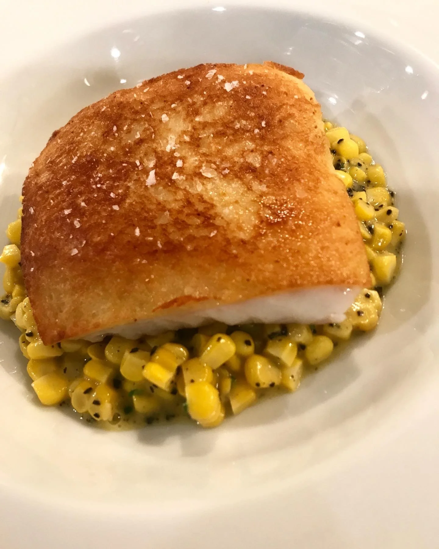 Baked fish fillet with golden-brown crispy skin, served over seasoned corn kernels in a white dish.