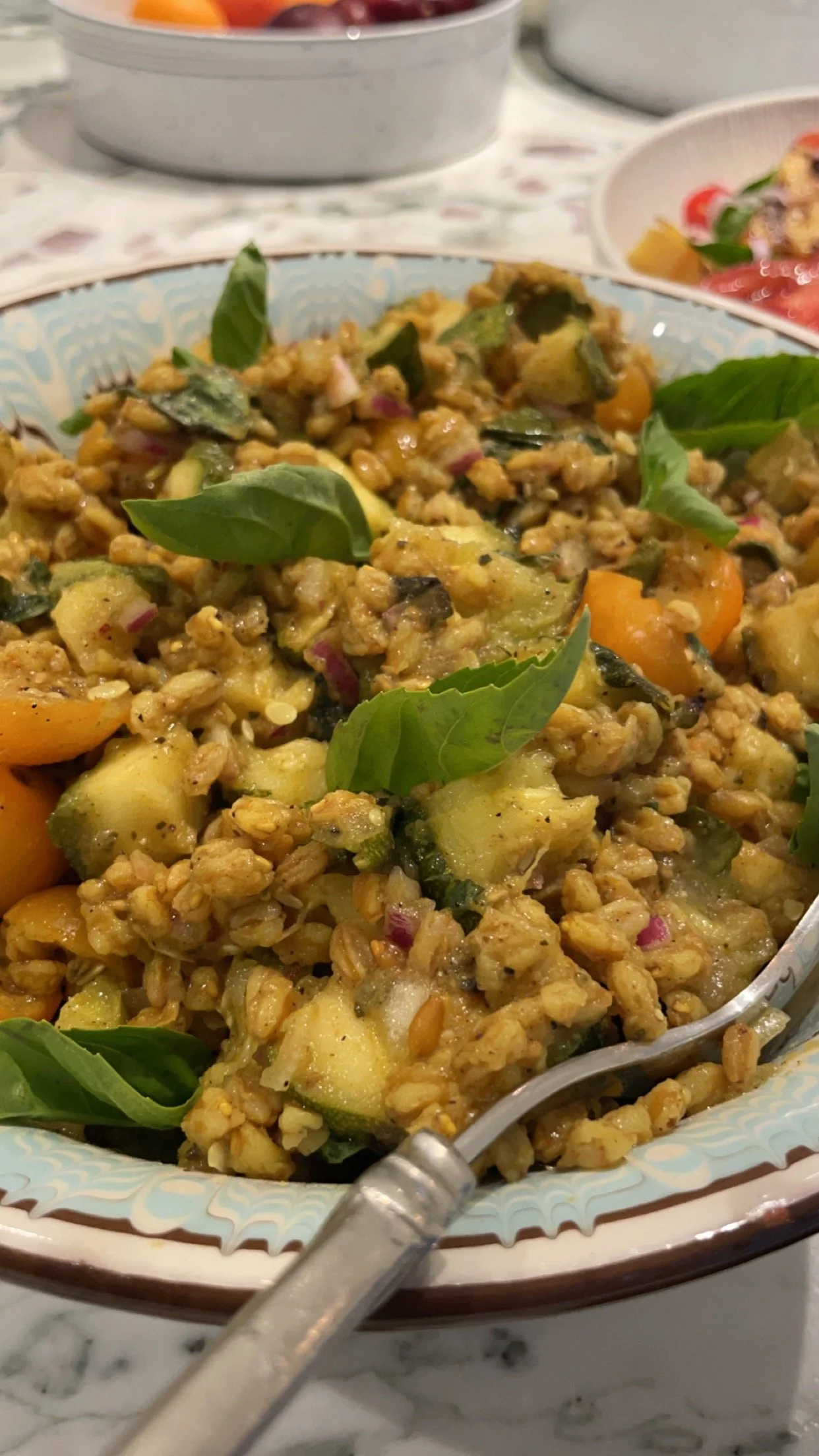 A bowl of mixed vegetable and grain salad garnished with fresh basil leaves on a patterned plate.