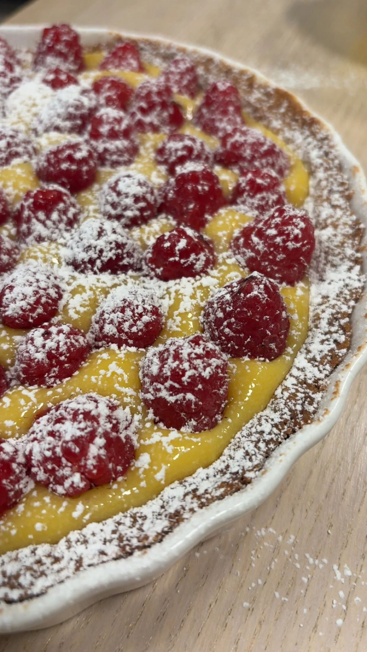 Close-up of a dessert pie with raspberries, lemon filling, powdered sugar, and a crust in a white dish.