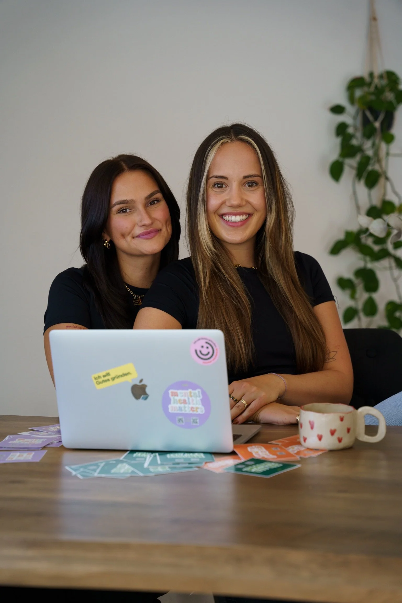 Two women sit at a table with a laptop and a mug with hearts, smiling at the camera in a room with a white wall and green plant in the background.