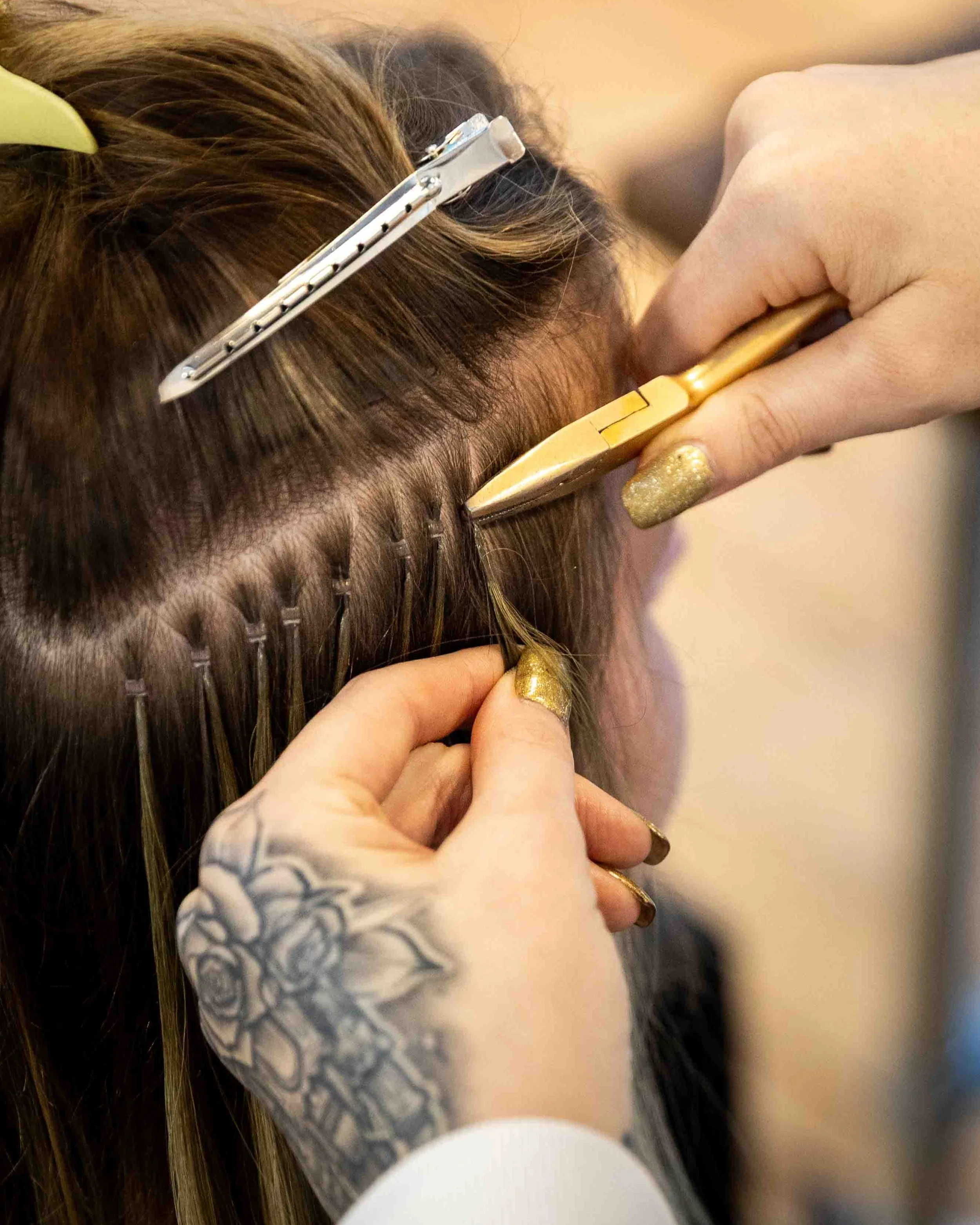 A hair stylist holding hair with one hand and weaving hair extensions with the other, while a hair technician installs hair extensions