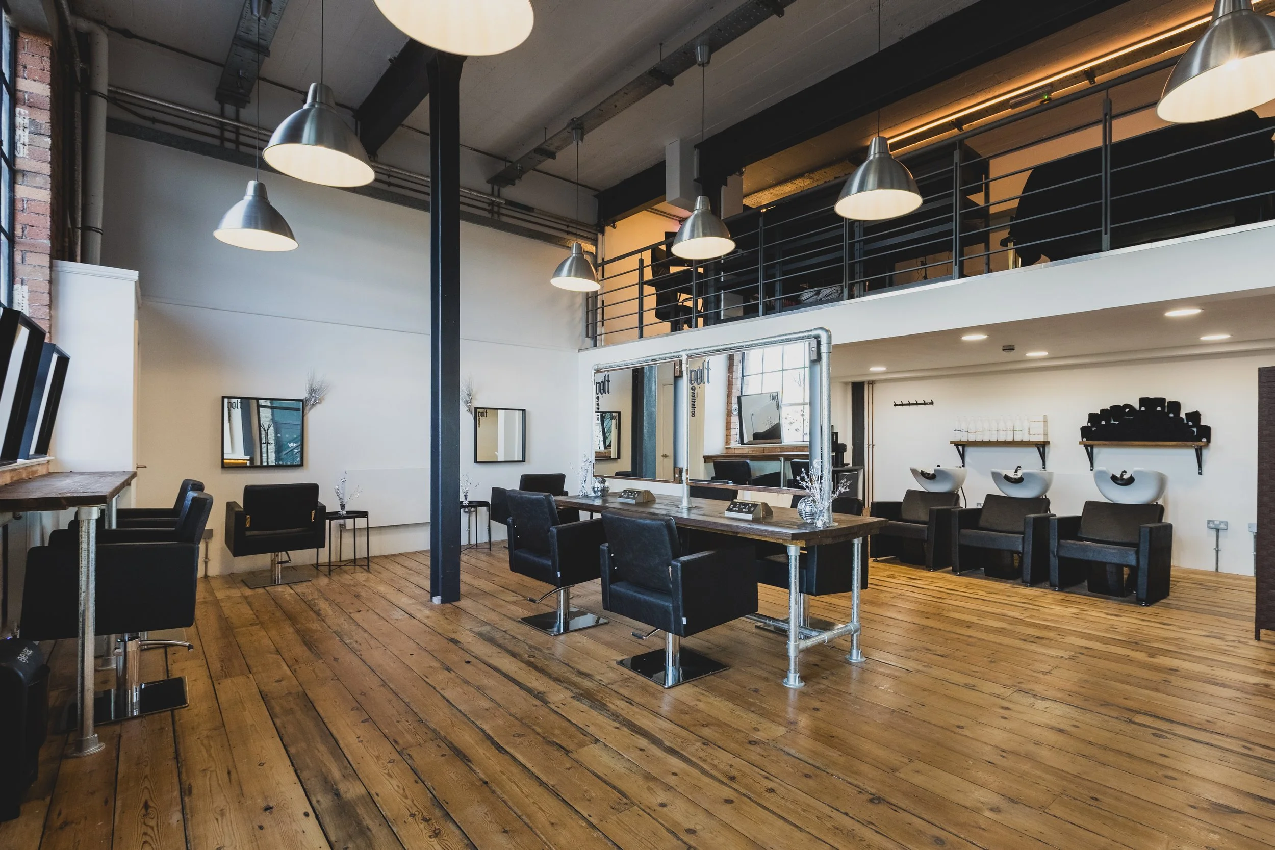 Interior of a modern hair salon in bristol with wooden flooring, black chairs, mirrors on the white walls, and hair washing stations in the background, with a loft area overhead.