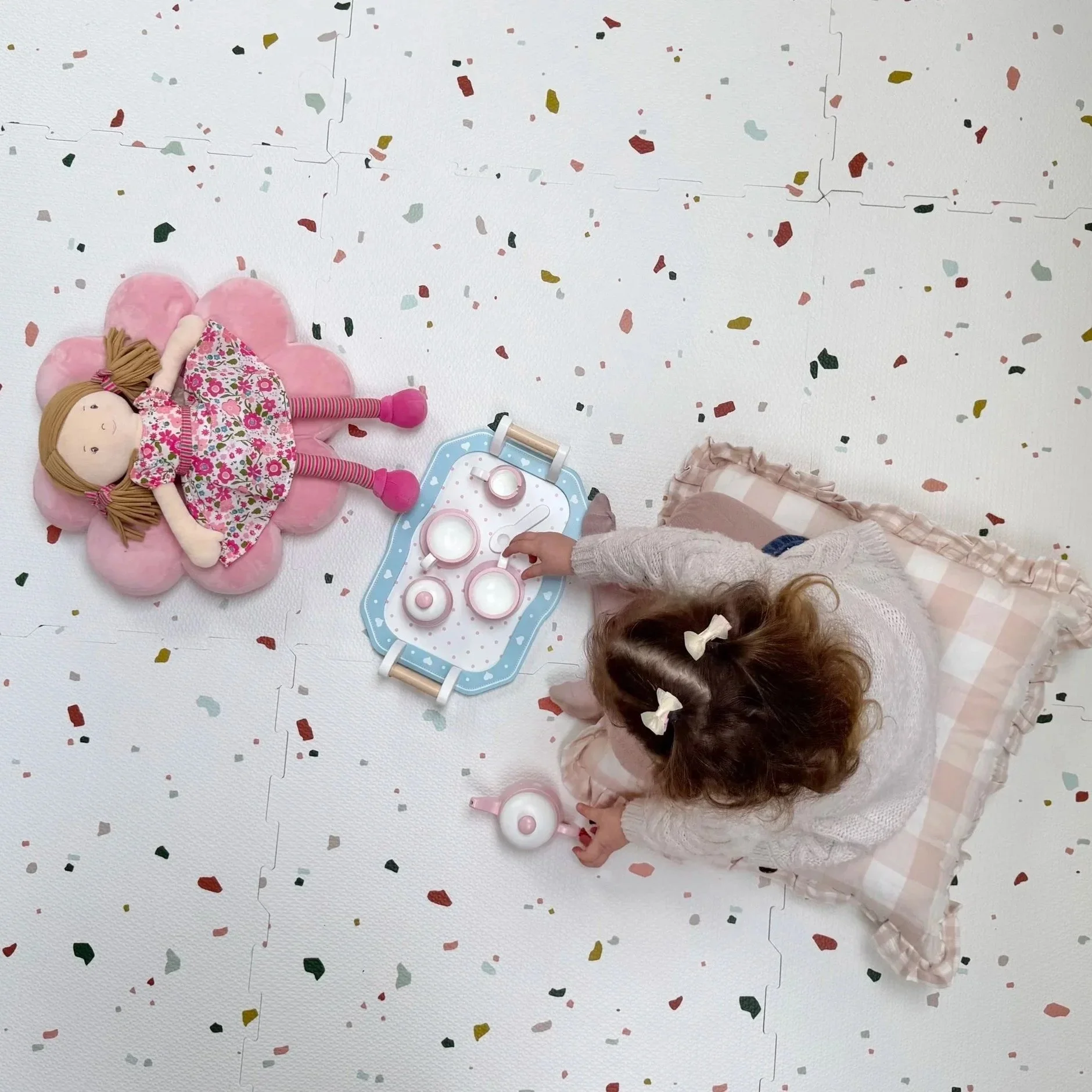 A young girl with curly hair and hair bows playing with a toy tea set and a doll on a pink flower-shaped cushion on a white confetti-patterned floor.