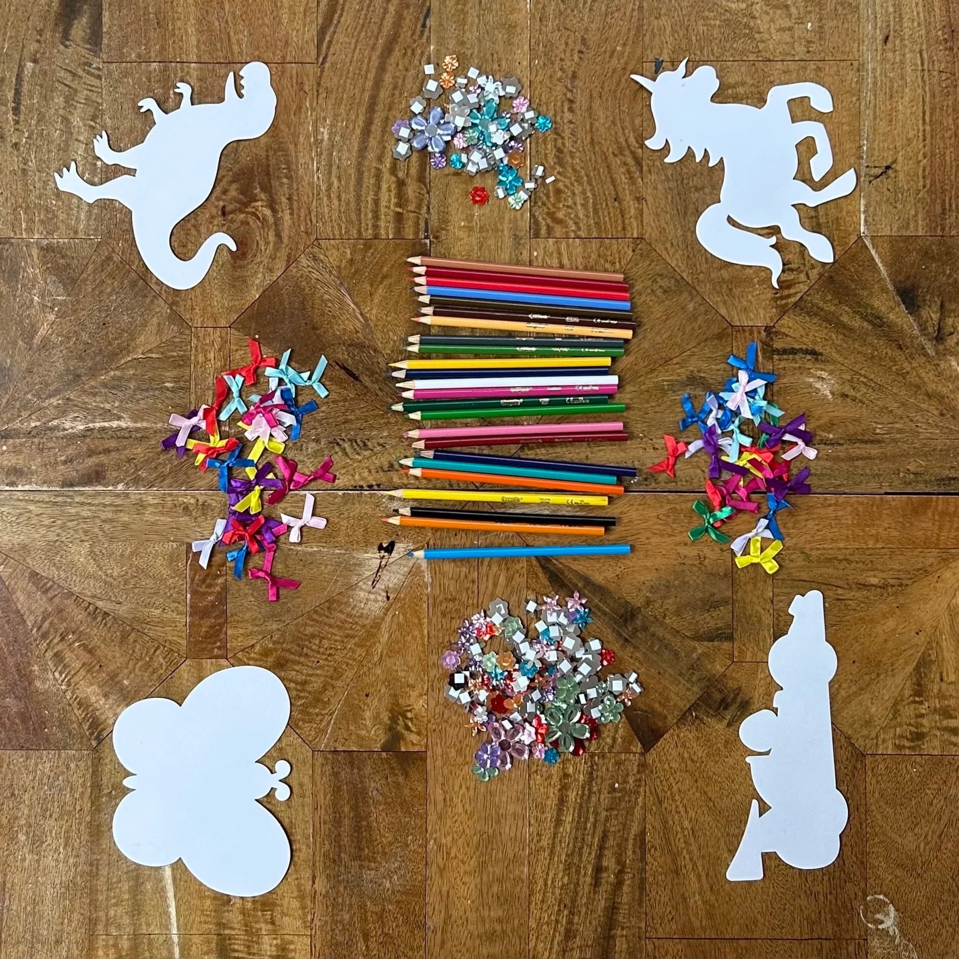 Decorative craft supplies including paper cutouts of dinosaurs, unicorns, and a bear, colorful pencils, and assorted sequins and ribbons arranged on a wooden surface.