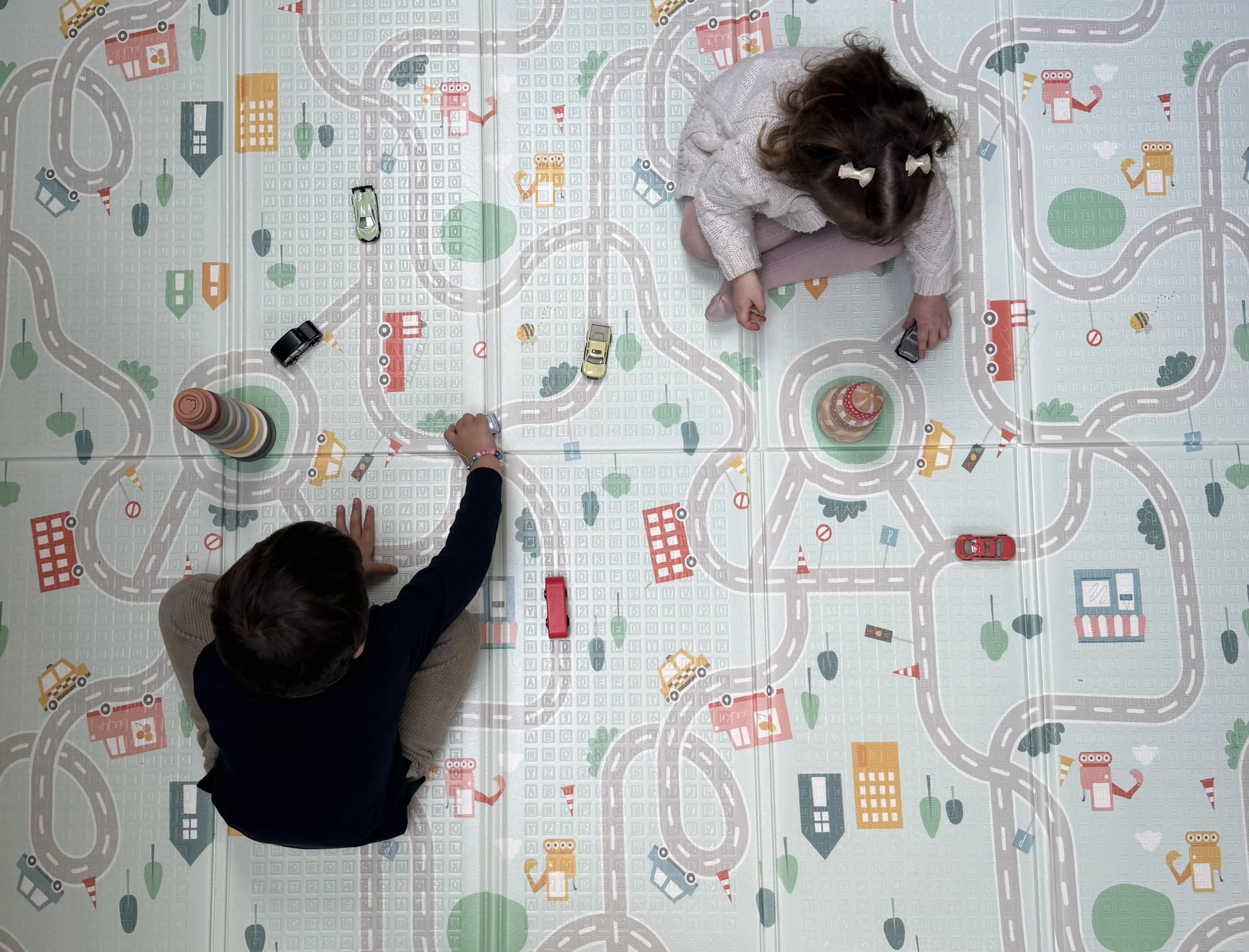 Two children playing on a colorful city-themed playmat with toy cars, buildings, and roads, one girl and one boy, viewed from above.