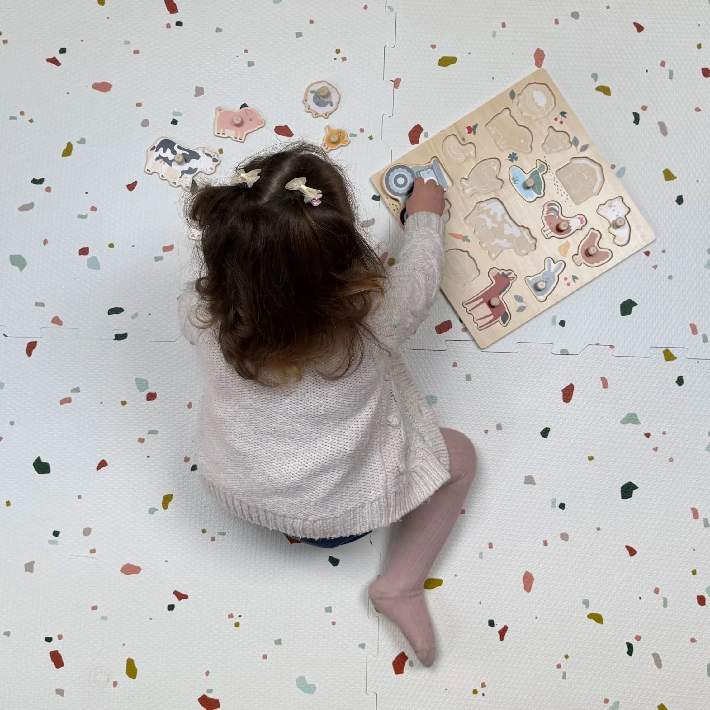 A young girl with curly hair and hair clips, sitting on a terrazzo floor and playing with animal-shaped puzzle pieces and toys.