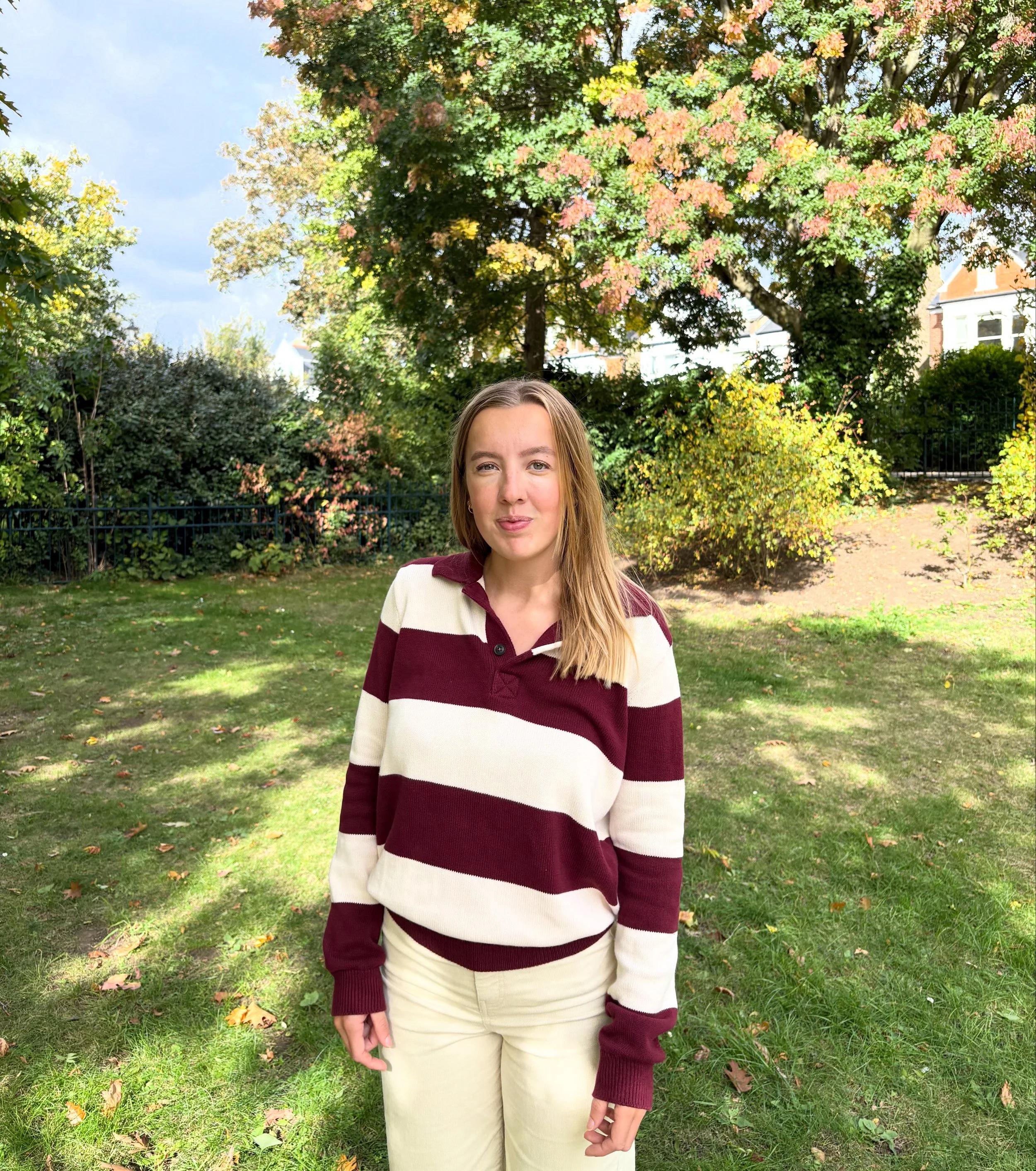 A young woman with shoulder-length light brown hair standing outdoors in a park with lush trees and grass, wearing a maroon and white striped long-sleeve shirt and light-colored pants.
