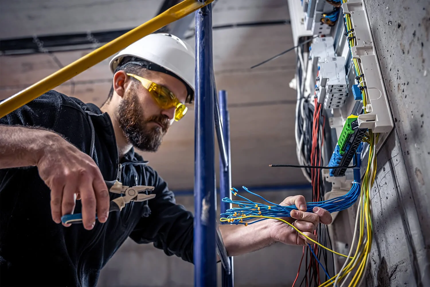 A technician working on electrical wiring, wearing safety glasses, a white safety helmet, and a black shirt, with organized wires and electrical components on a panel.