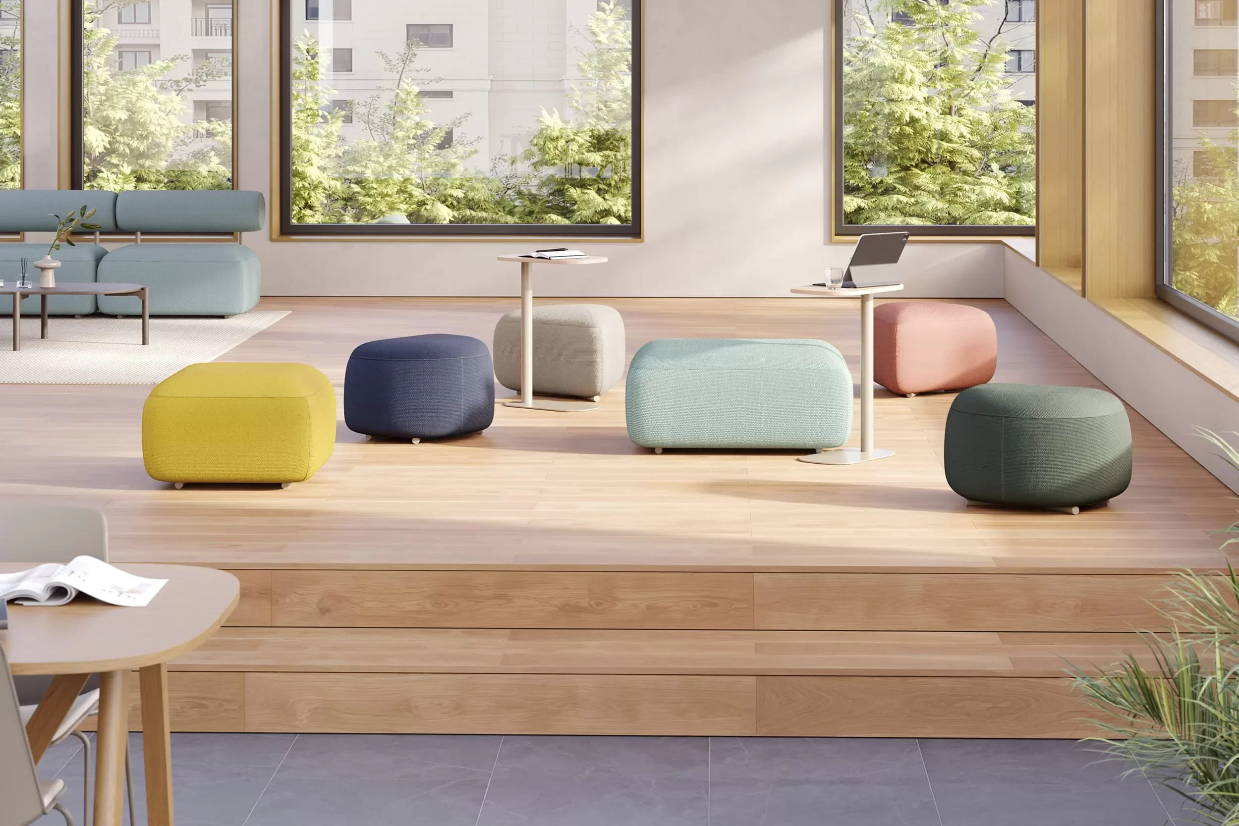 A bright, modern lounge area with various colorful ottomans, small side tables, a sofa, large windows with a view of trees and buildings, and hardwood flooring.