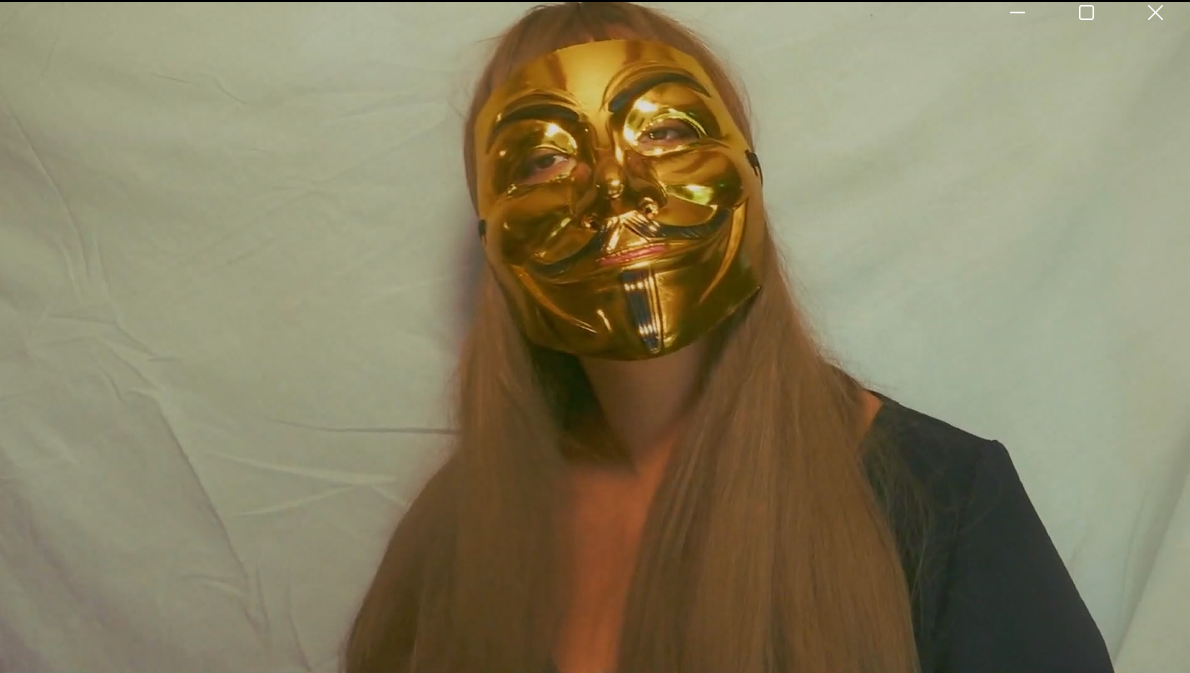Person with long hair wearing a shiny gold Guy Fawkes mask, standing against a plain background.