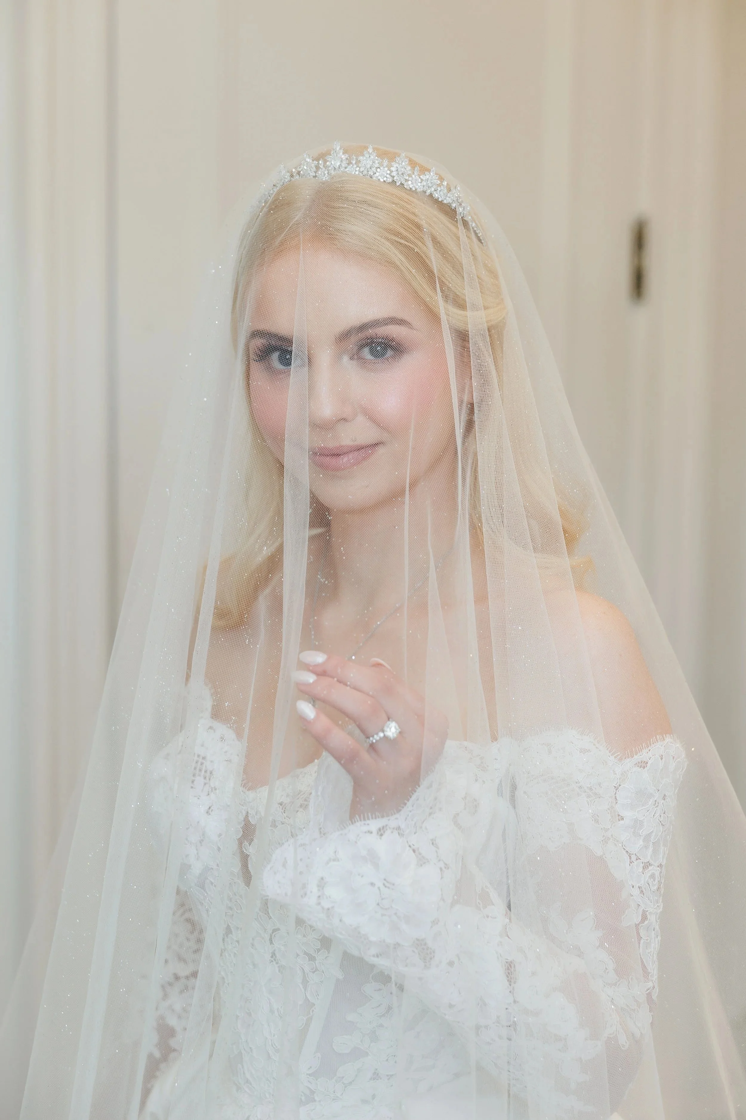 A bride with blonde hair in a lace wedding dress, wearing a tiara and veil, smiling at the camera, holding her hand with a diamond ring near her face.