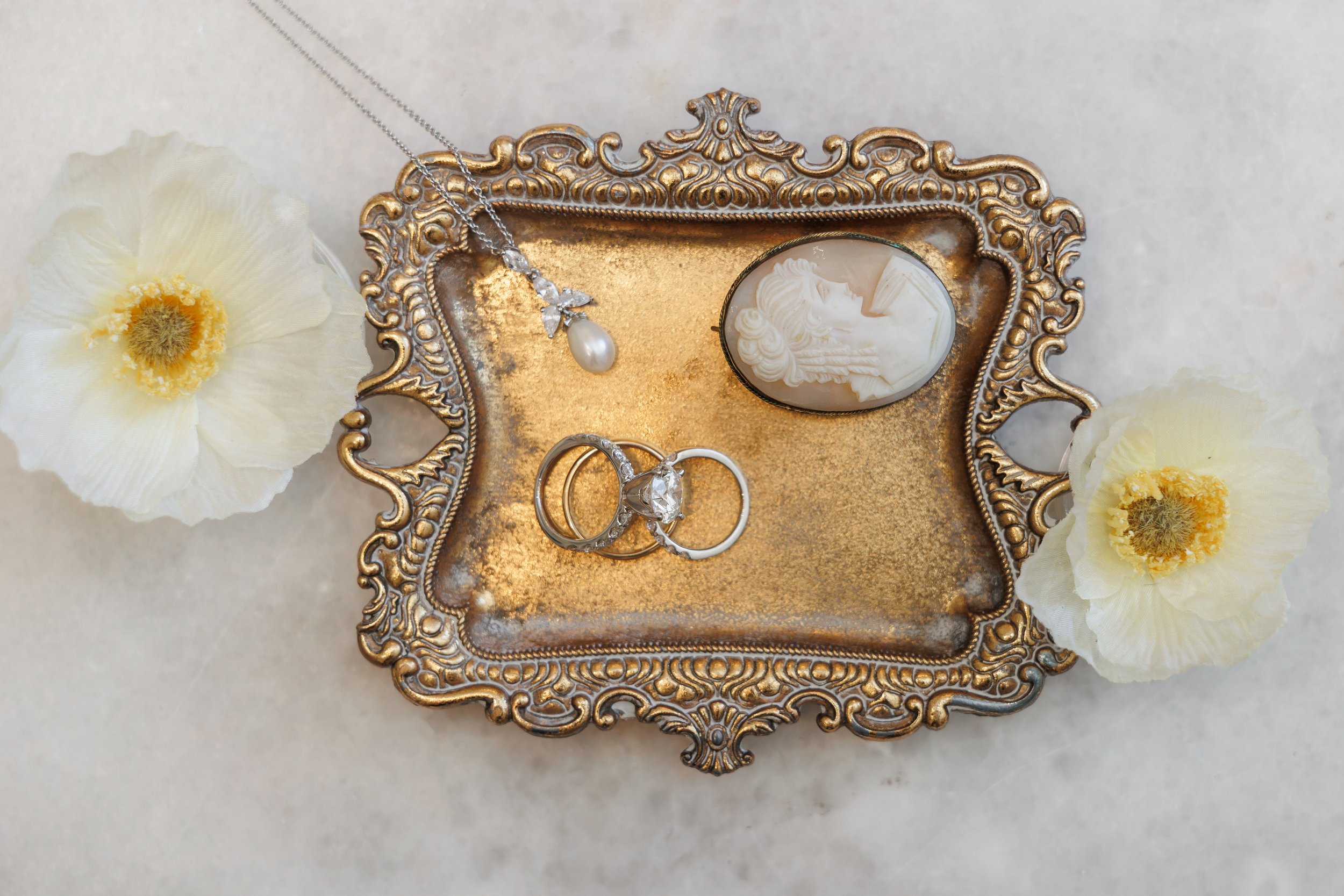 An ornate gold tray holding jewelry, including a silver necklace with a pearl pendant, a silver ring with a large gemstone, and another ring with small stones. The tray is decorated with white flowers on a light background.