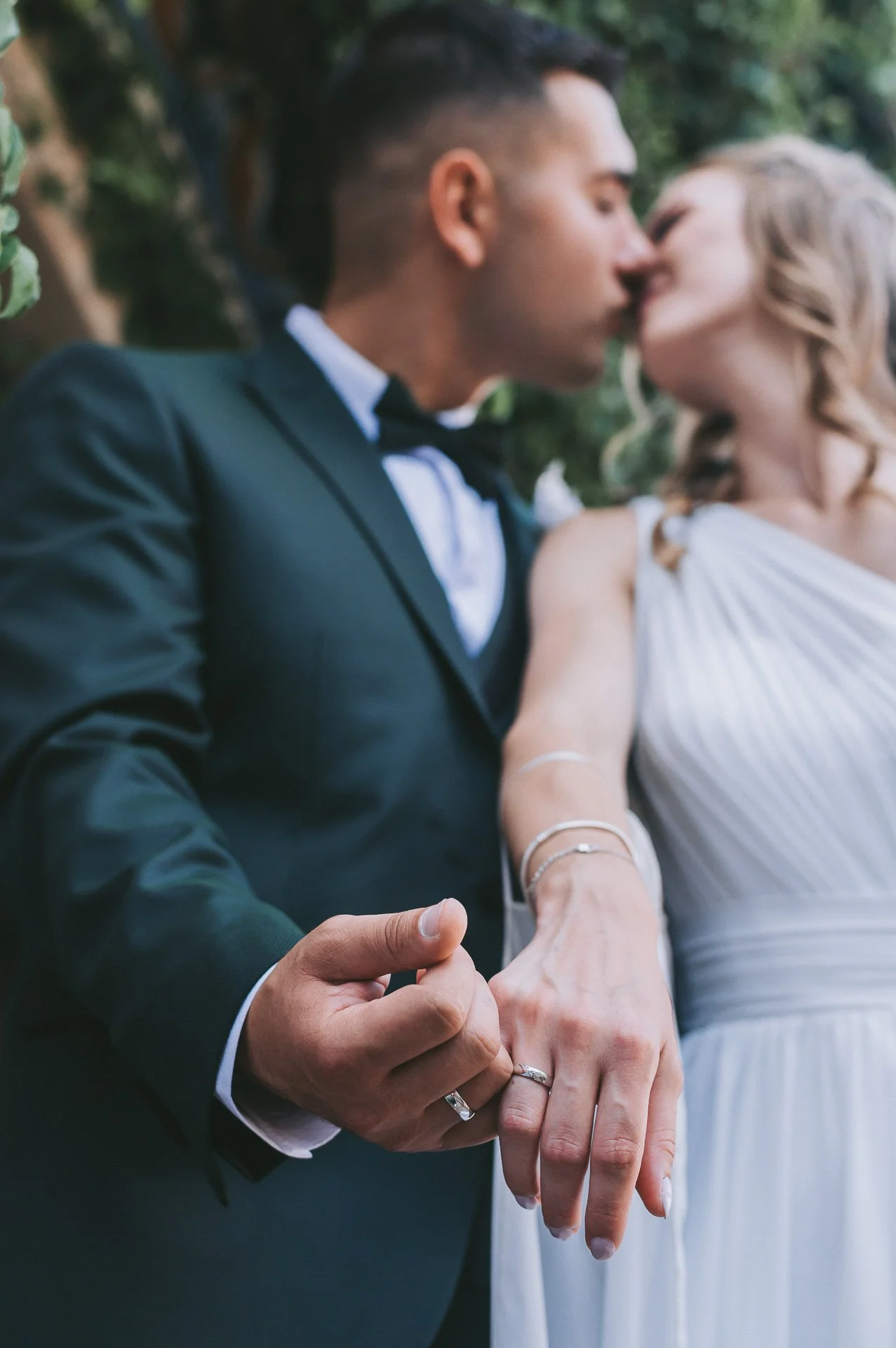 A bride and groom kissing outdoors, holding hands and showing their wedding rings.