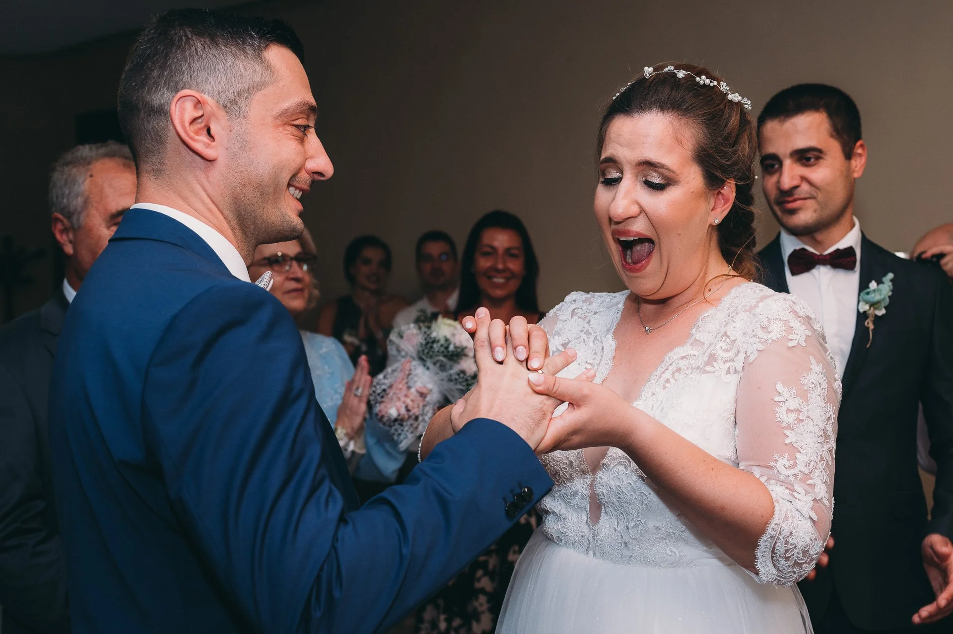 A bride and groom exchanging rings during their wedding ceremony, with friends and family in the background.