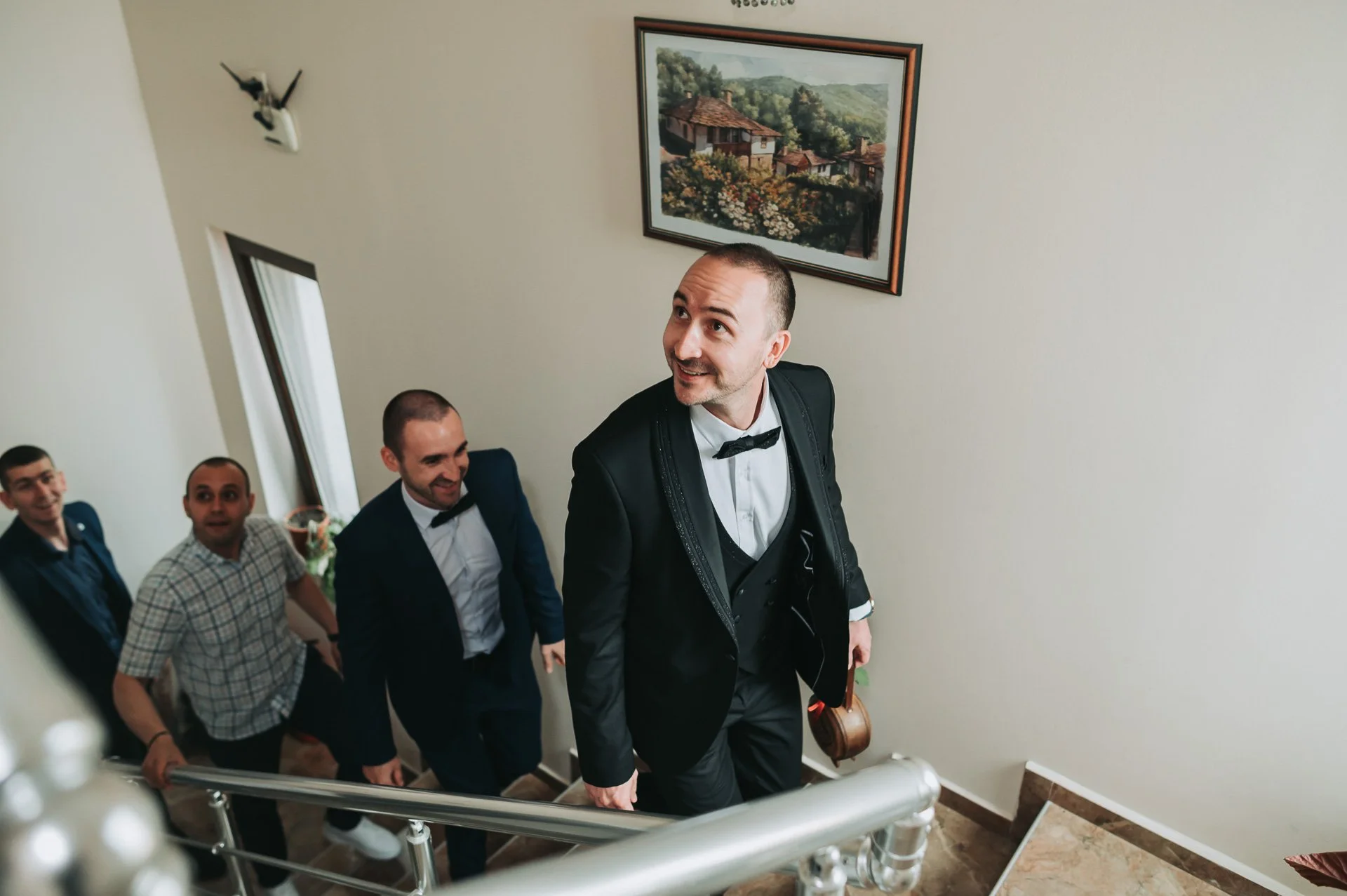A man in a tuxedo with a bow tie, holding a small brown bag, leading a group of men up a staircase, smiling. The staircase has a metallic railing, and there is a framed painting of a landscape on the wall behind them.