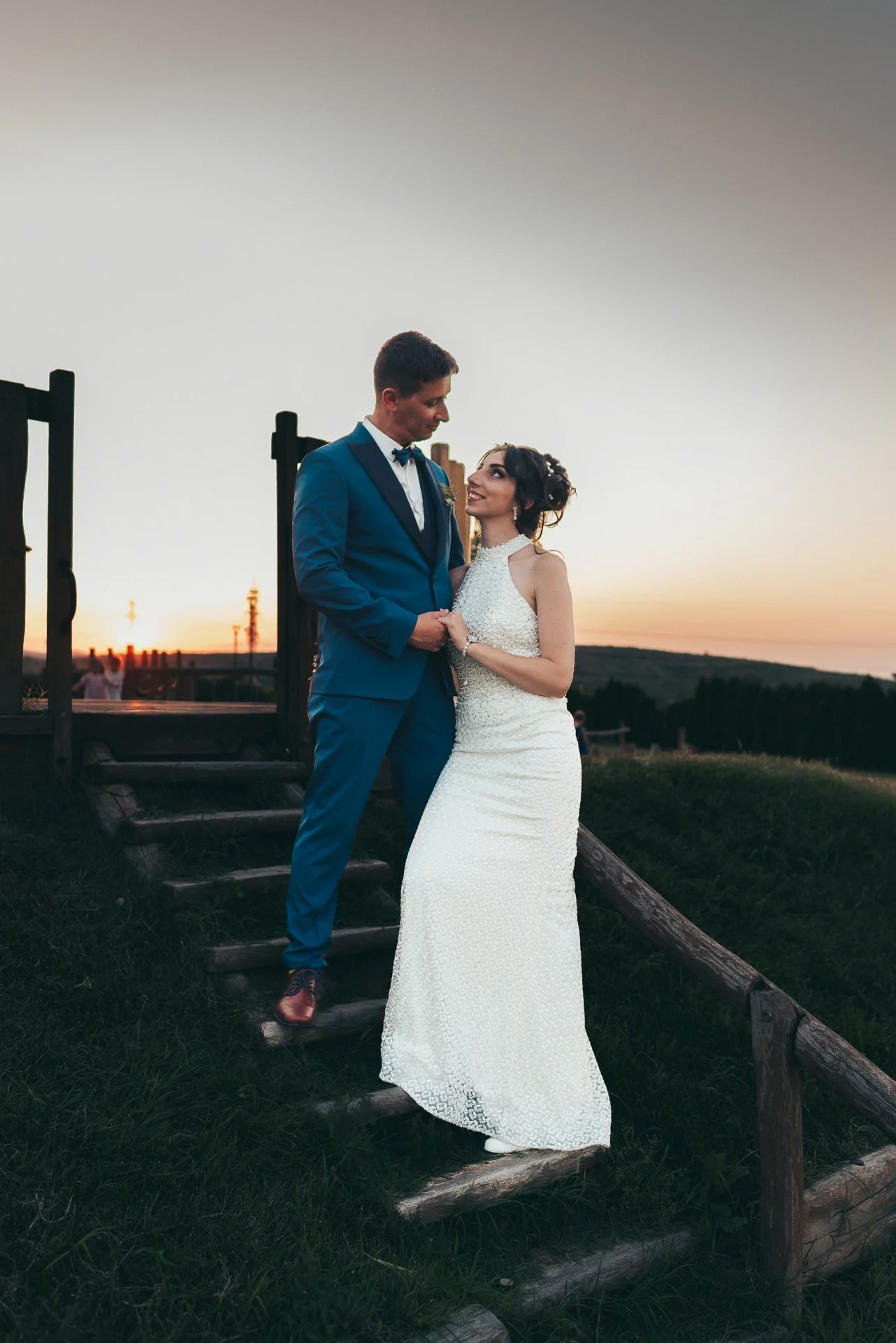 A bride and groom standing on a wooden staircase outdoors at sunset, gazing at each other, with the sunset and a distant landscape in the background.
