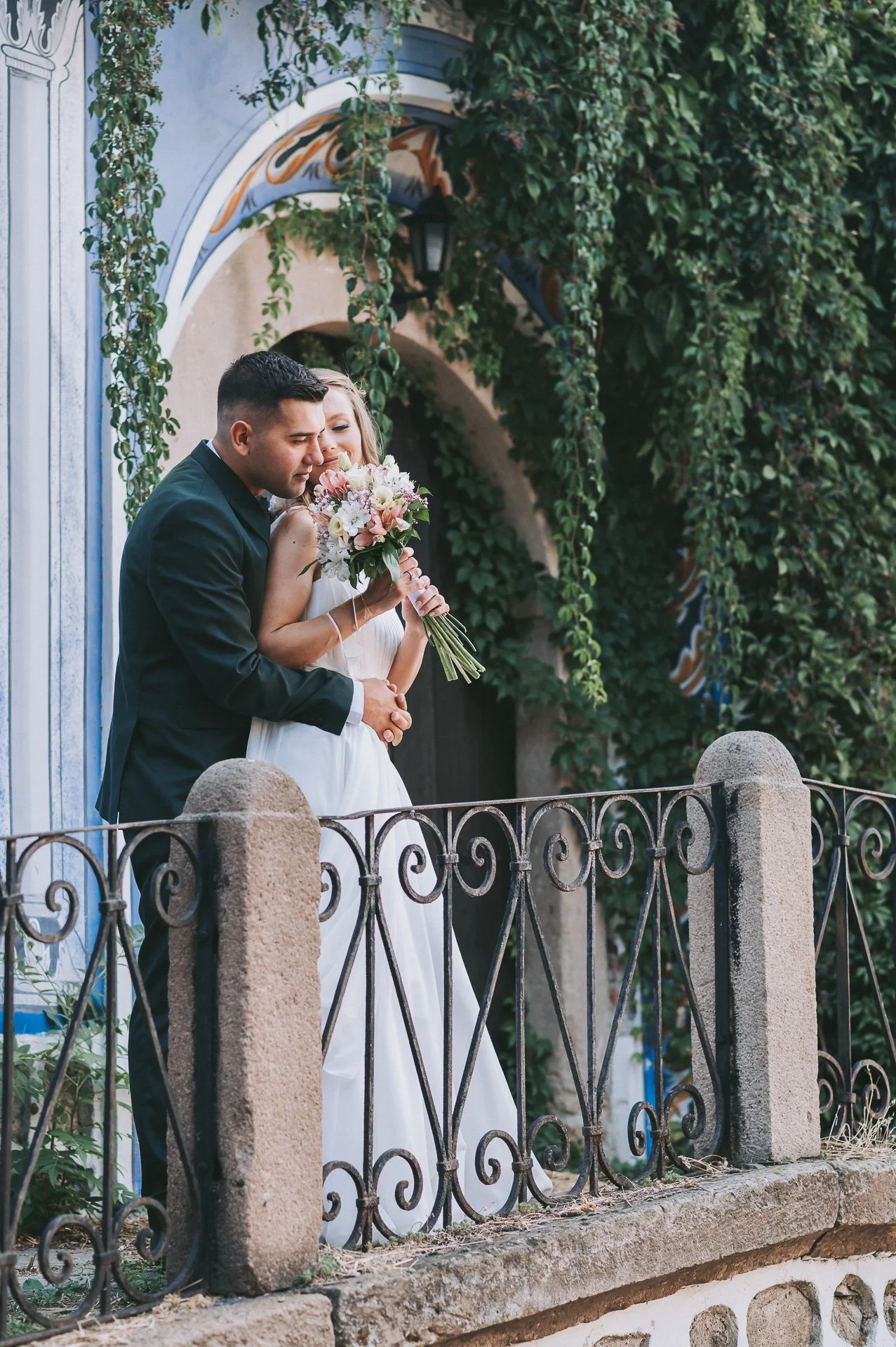 Couple on a balcony, with the woman holding a bouquet of flowers and leaning into the man, surrounded by greenery and a decorative metal railing.