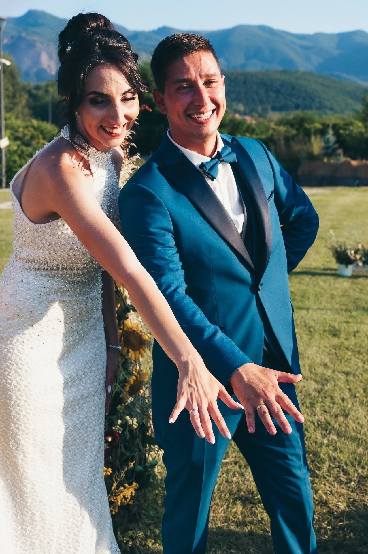 A bride and groom at their wedding, smiling and showing their wedding rings, outdoors with mountains and greenery in the background.