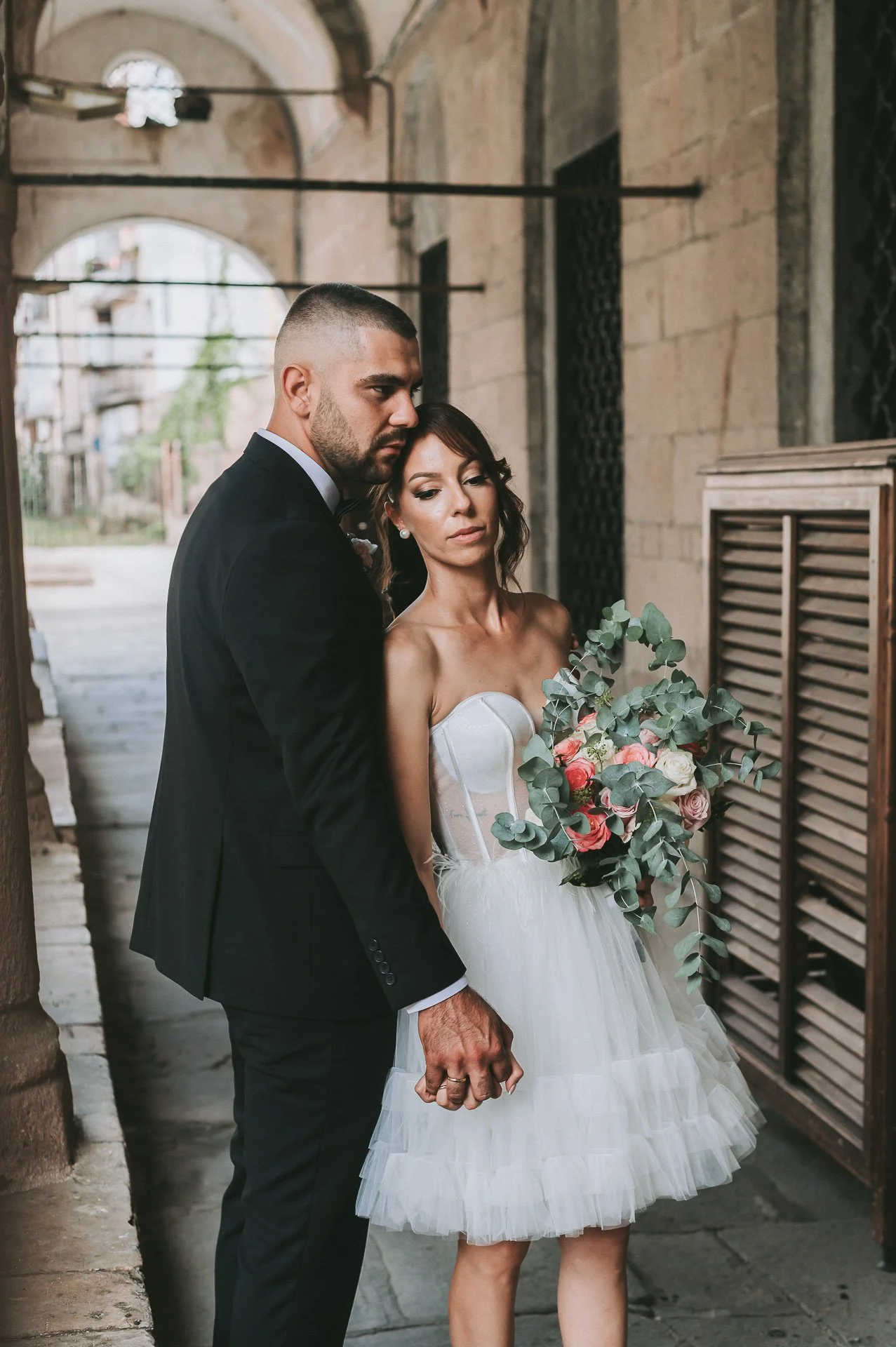 A bride and groom standing close together, holding hands, outside a building with stone walls and archways. The bride wears a strapless white dress and holds a bouquet of pink and white flowers with green eucalyptus. The groom wears a black suit and 