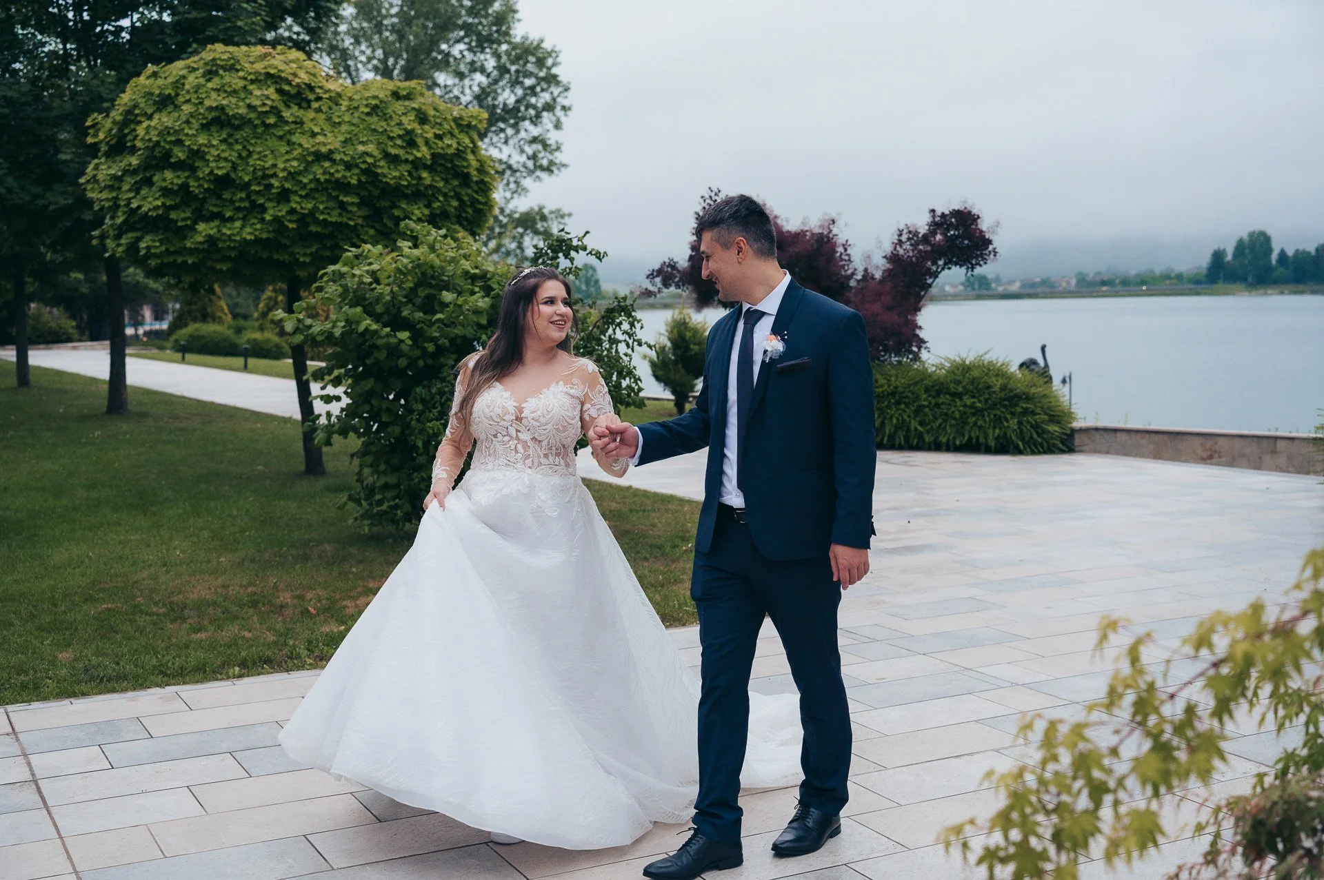 A bride and groom holding hands and smiling at each other outside near a lake, with trees and shrubs around, on a cloudy day.
