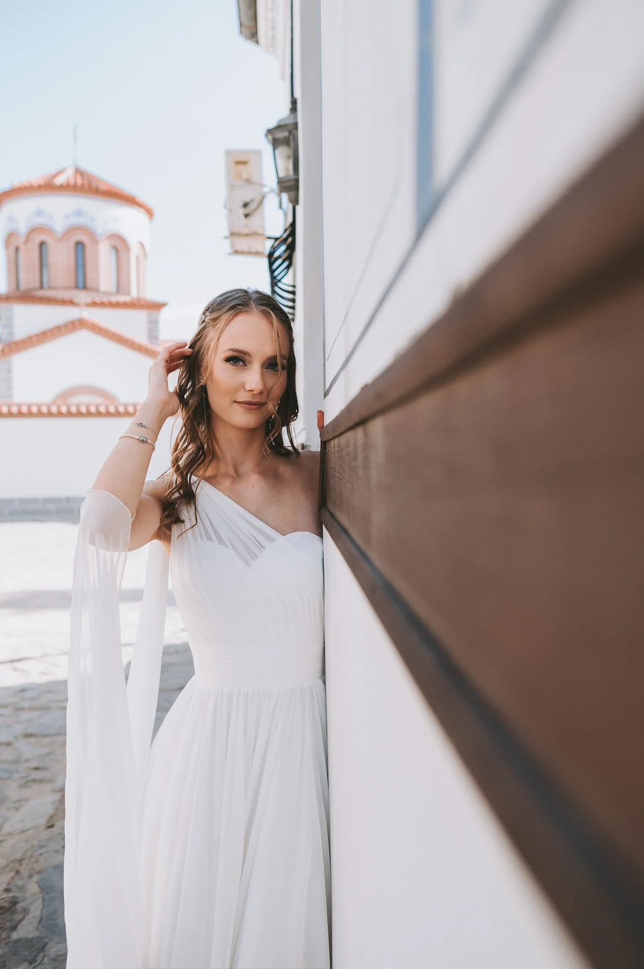 A woman in a white dress standing next to a building with her hand touching her hair, with a church in the background.