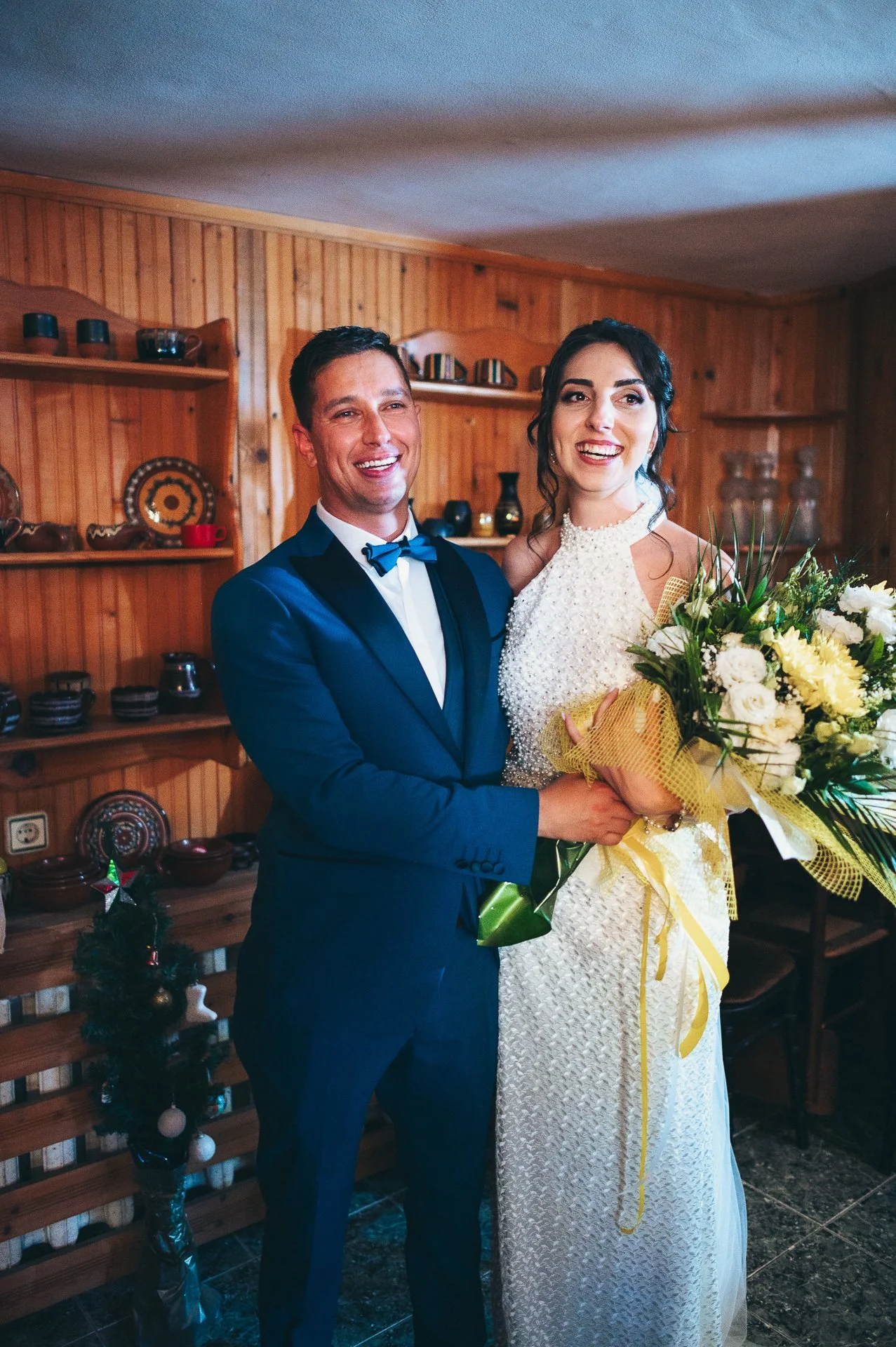 A couple dressed in wedding attire, standing indoors, smiling, with the man in a blue suit and bow tie and the woman in a white wedding dress holding a bouquet of flowers.