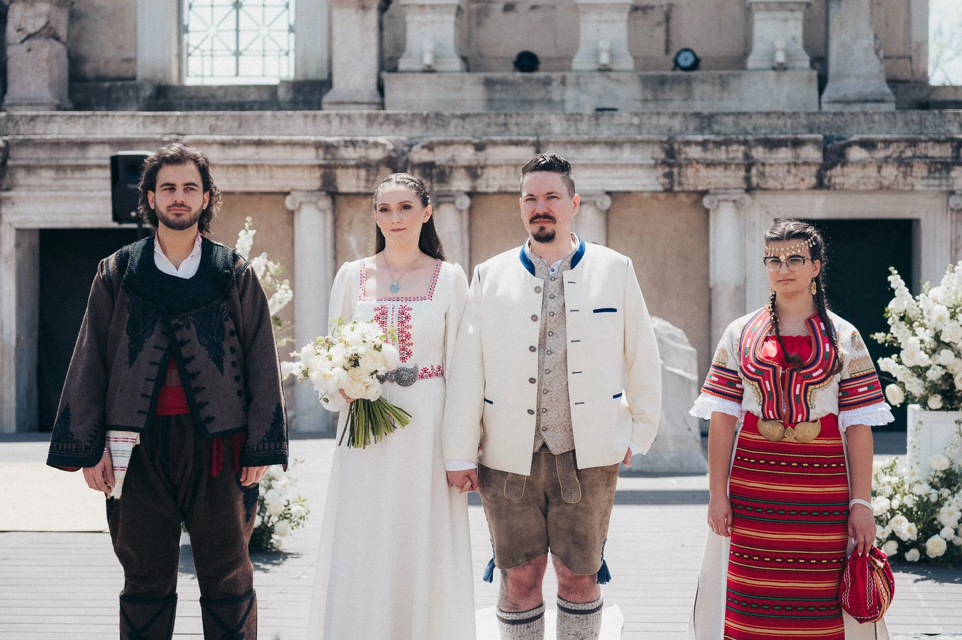 Group of four people standing outdoors, dressed in traditional Eastern European (possibly Balkan) traditional clothing, in front of a historic stone building with columns, during daytime.