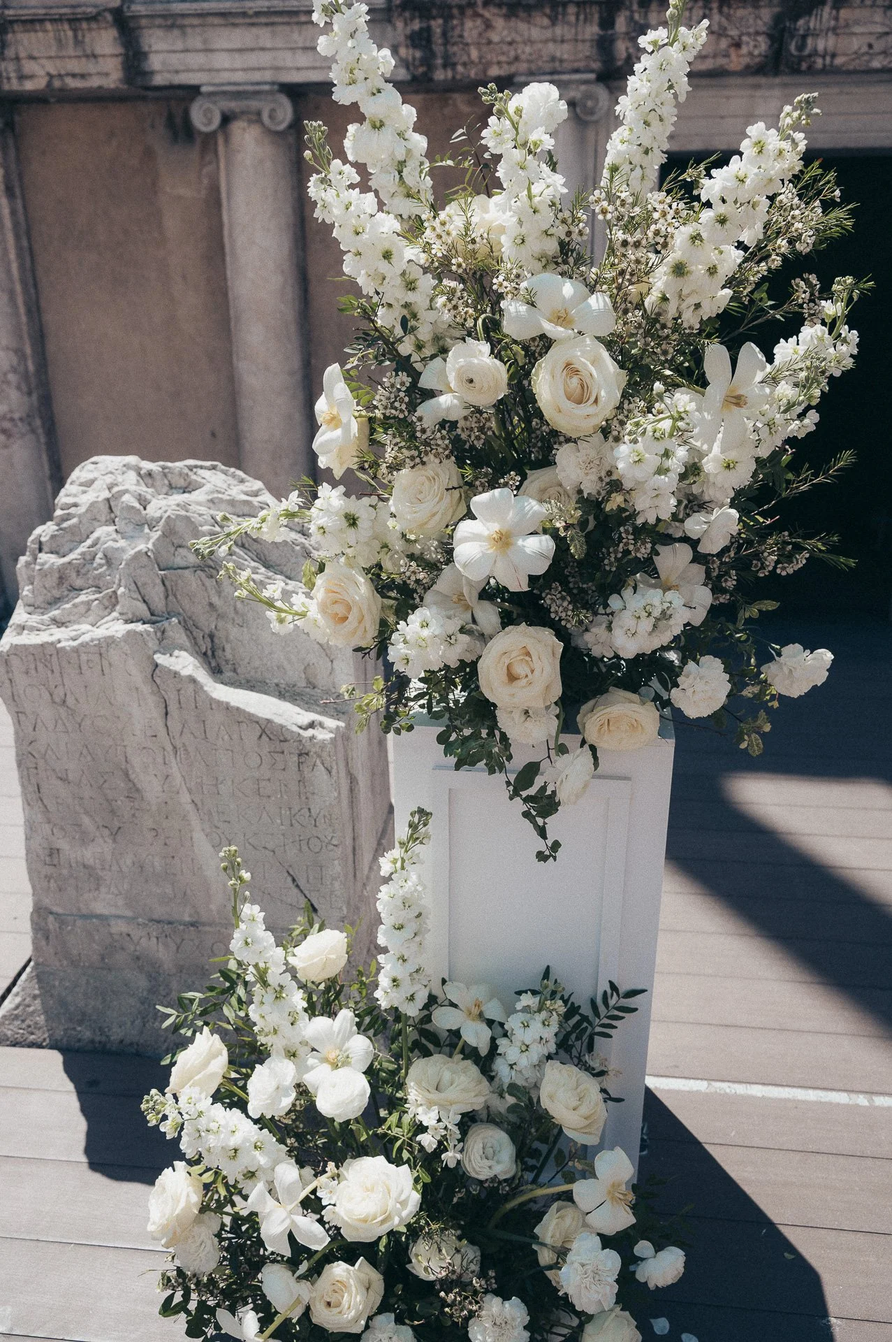 A large white flower arrangement with roses, orchids, and other white flowers in a white rectangular vase on a wooden floor, with a broken stone tablet with Greek inscriptions in the background.
