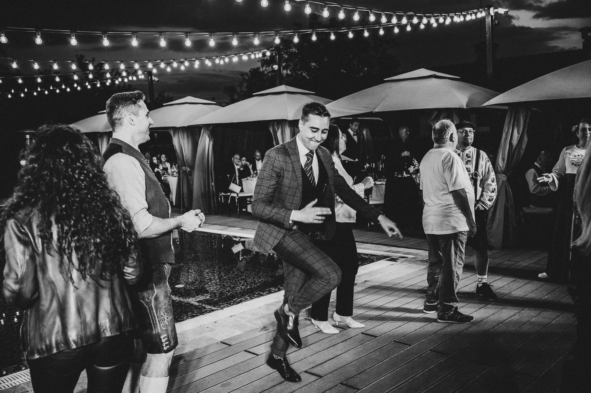People dancing outdoors at night under string lights, with tables and umbrellas in the background.