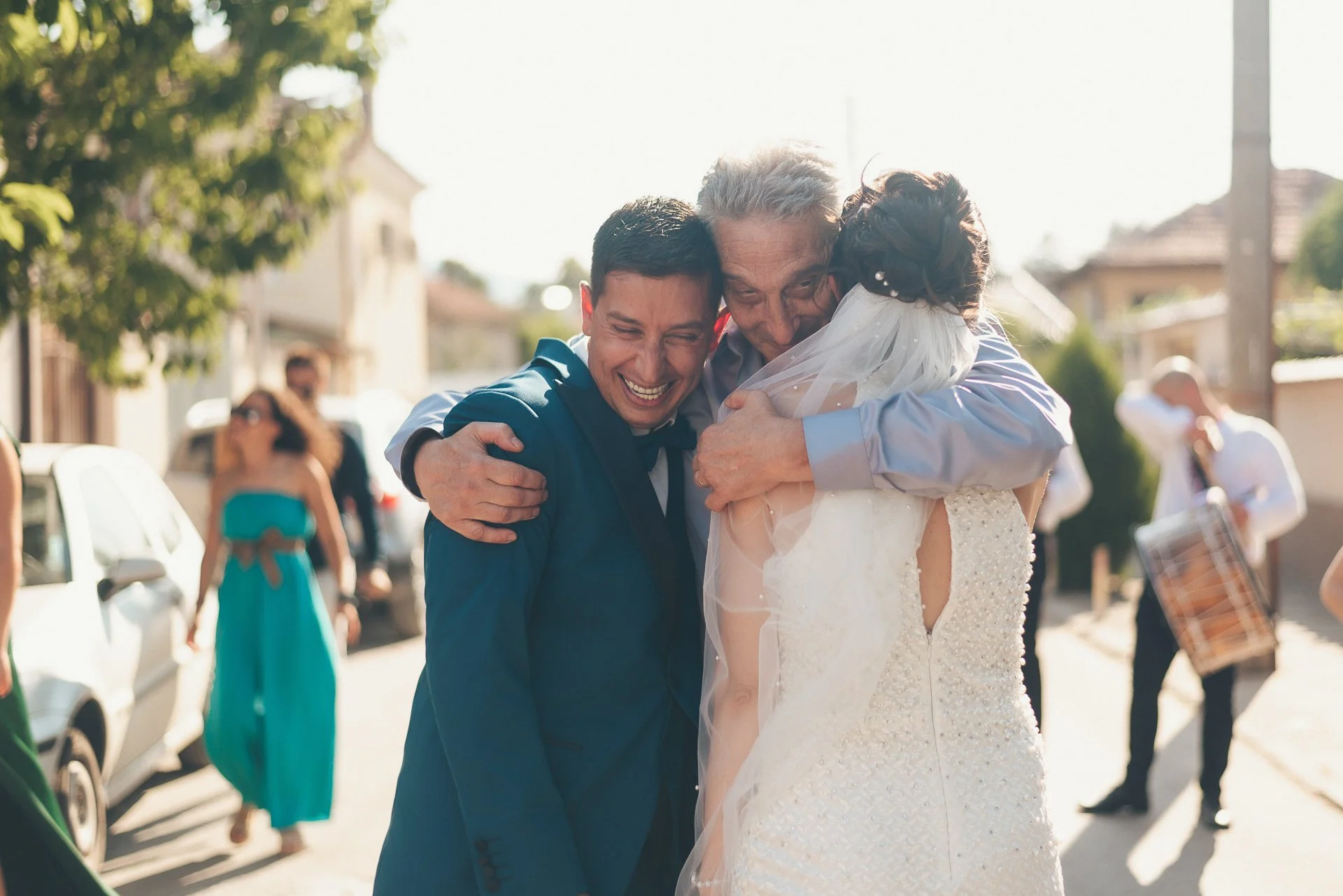 Family members hugging on a sunny street during a wedding celebration, with a bride in a white gown and veil.
