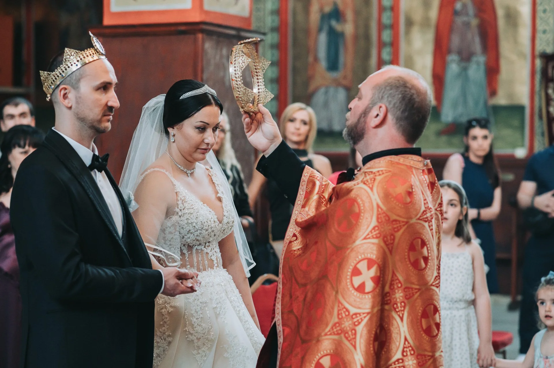 A priest performing a wedding ceremony in a church with the bride and groom standing before him. The priest is holding a religious object above the bride and groom. The bride wears a white wedding dress with lace details and a veil. The groom is dres