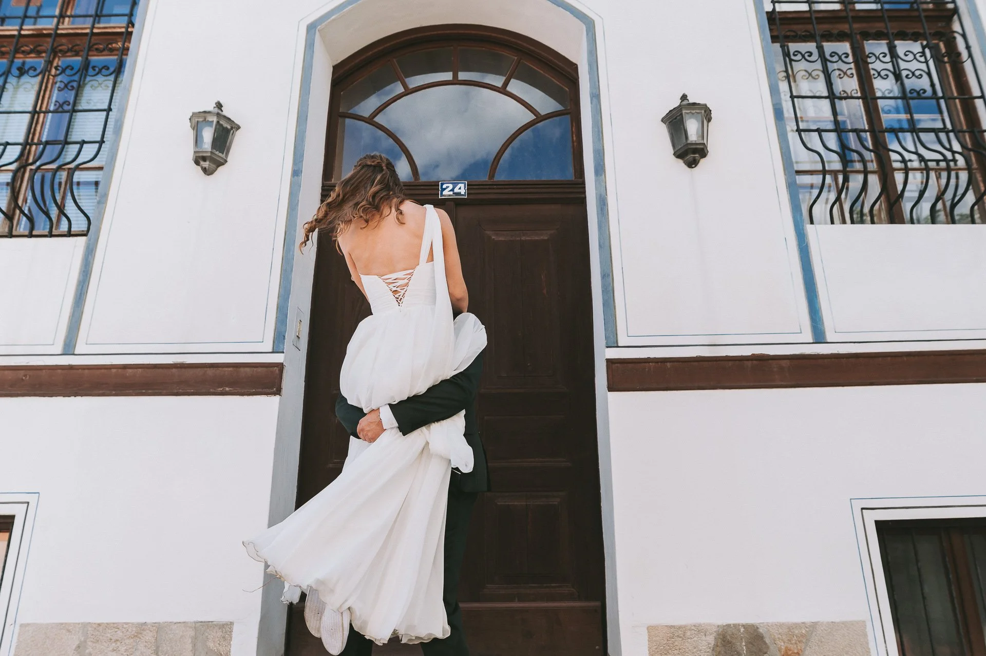 A couple in wedding attire, with the bride being carried by the groom, standing in front of a wooden door on a white building with windows and iron bars.