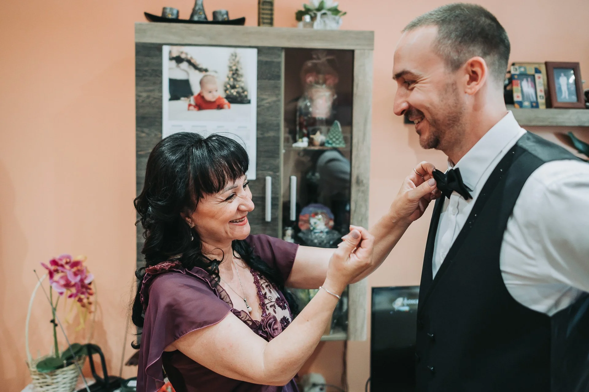 A woman helping a man adjust his black bow tie in a living room, both smiling. The woman has black hair and is wearing a purple dress. The man has light brown hair and is wearing a white shirt with a black vest and tie. Background includes a wooden c