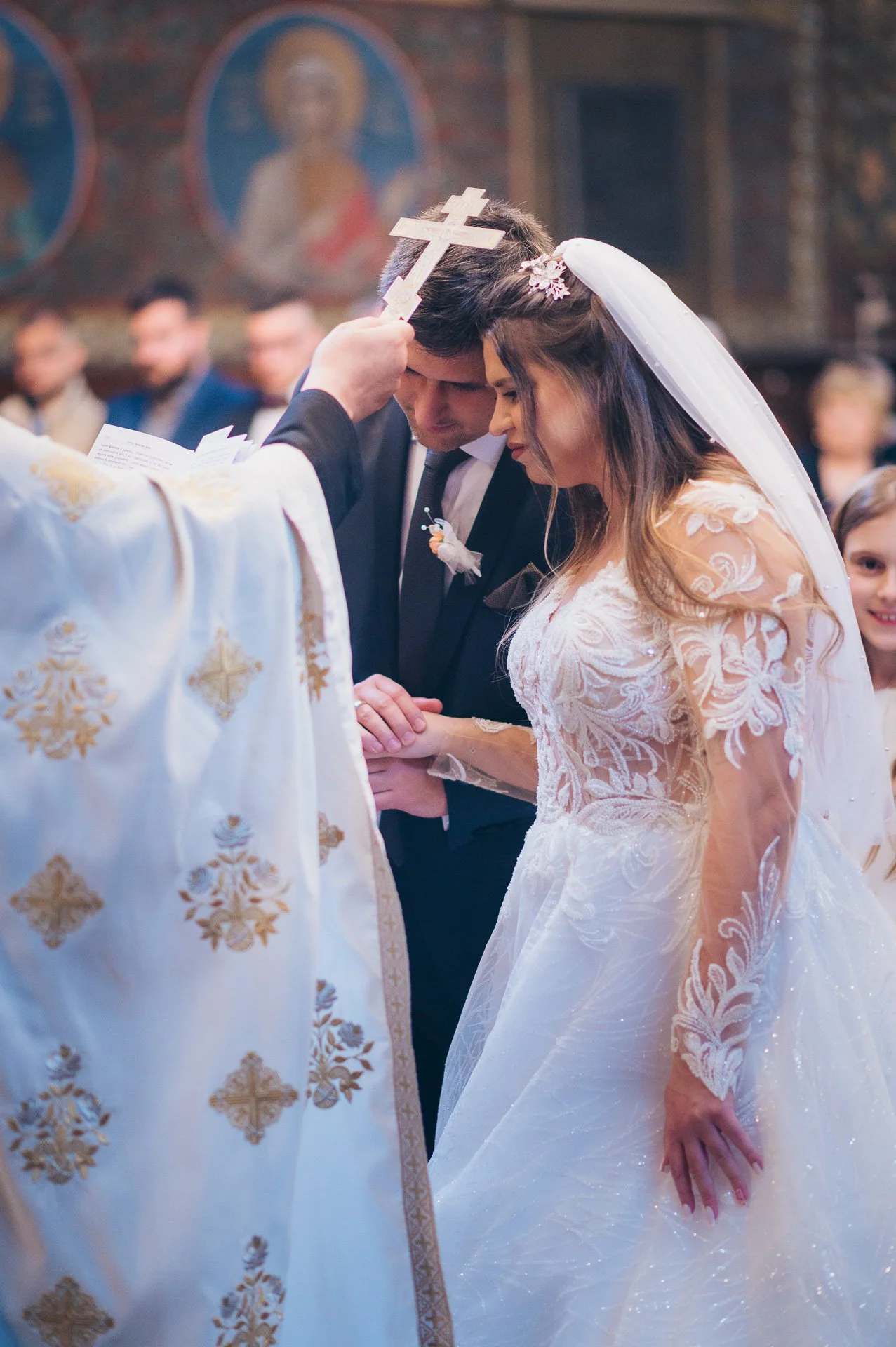 A bride and groom stand with heads bowed during a church wedding ceremony, with a priest holding a cross above them.