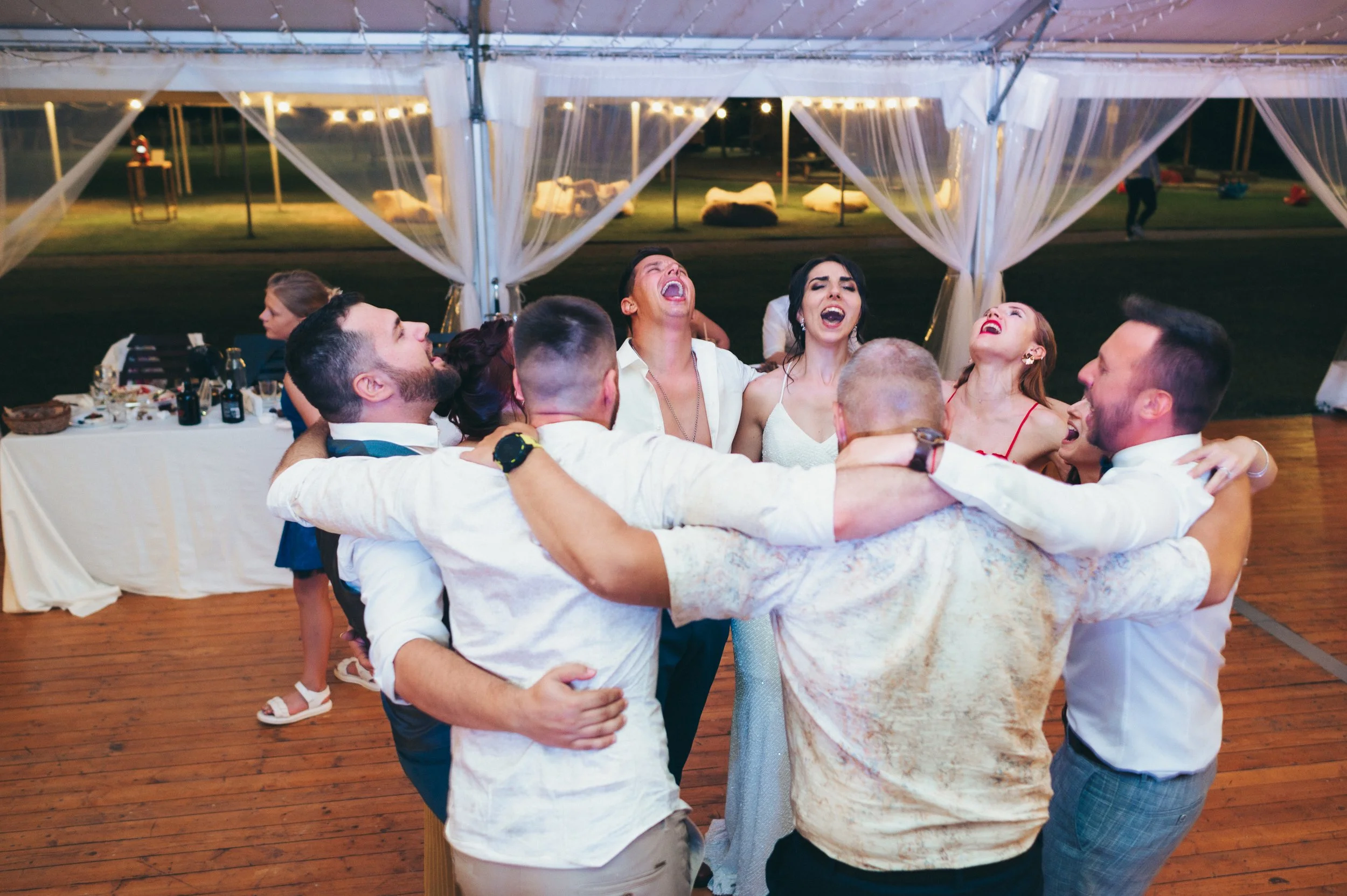 Group of people hugging and celebrating at a wedding reception or party under a tent, with smiles and cheerful expressions.