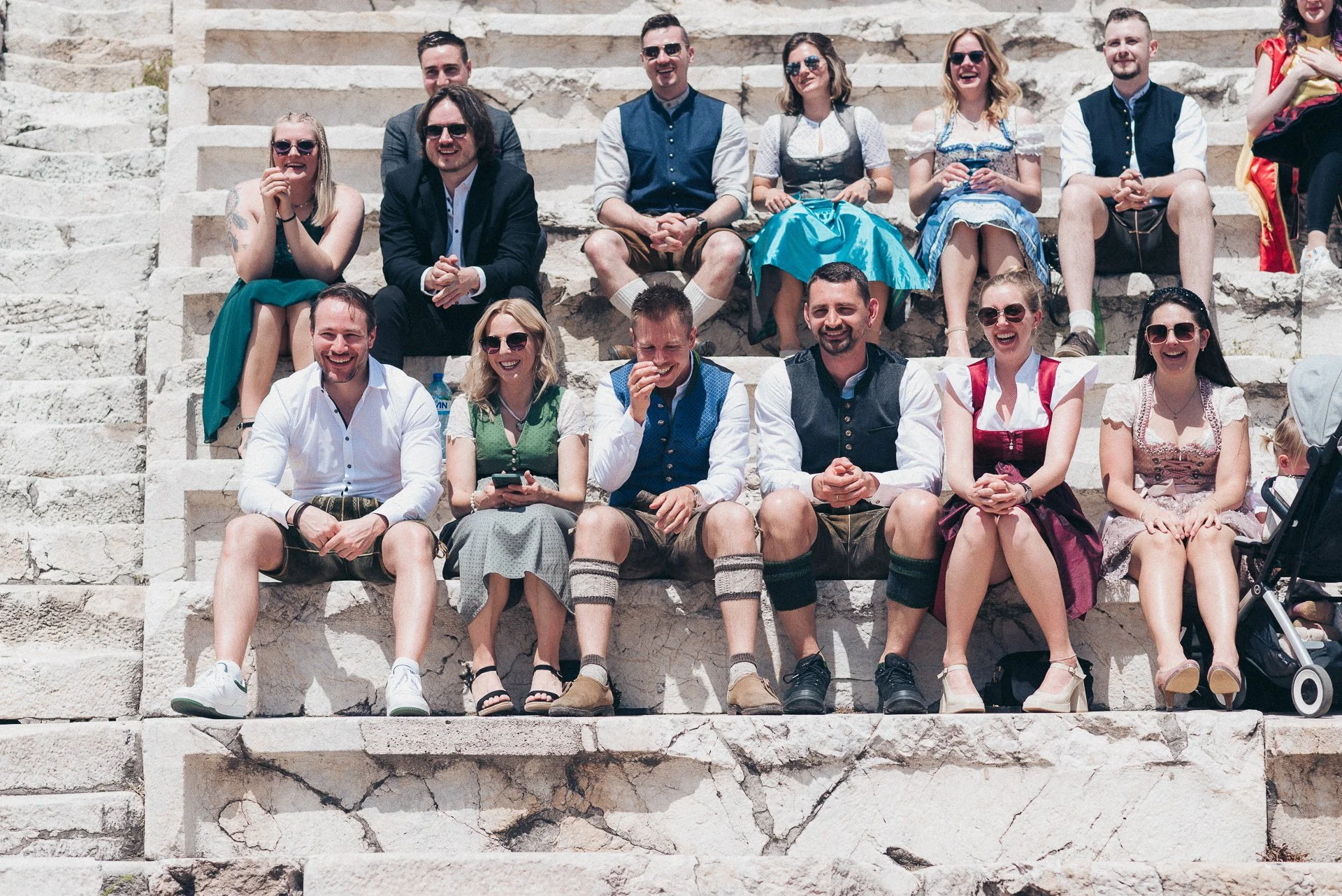 Group of people dressed in traditional Bavarian clothing sitting on stone steps outdoors, smiling and enjoying the day.
