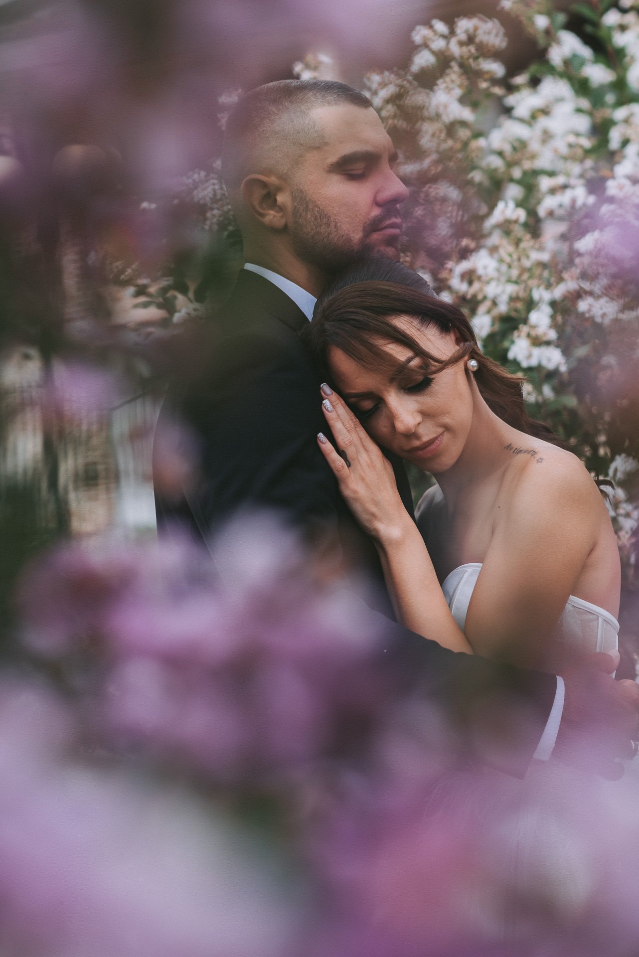 A man and woman embracing in a romantic moment surrounded by pink and white flowers, with the woman resting her head on the man's chest.