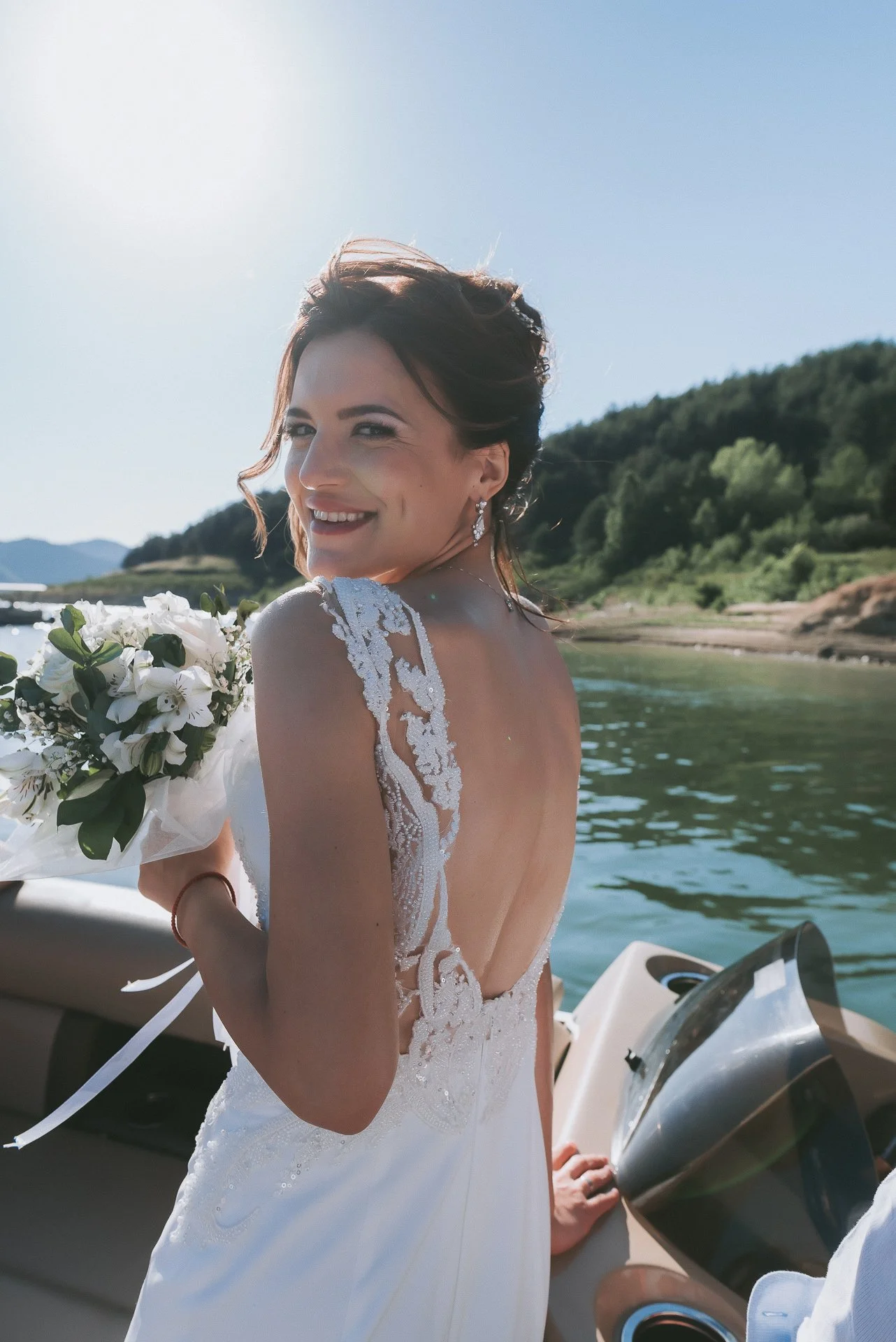 A bride in a wedding dress with lace details on the back and shoulders, holding a bouquet of white flowers, smiling on a boat near a body of water with green hills in the background.