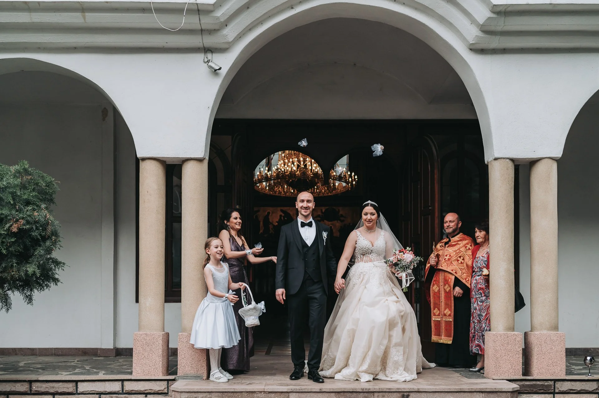 Wedding couple with family and friends outside a church, bride in a white gown with a bouquet, groom in a black tuxedo, celebrating with a smiling group of family members and a priest, in front of church entrance.