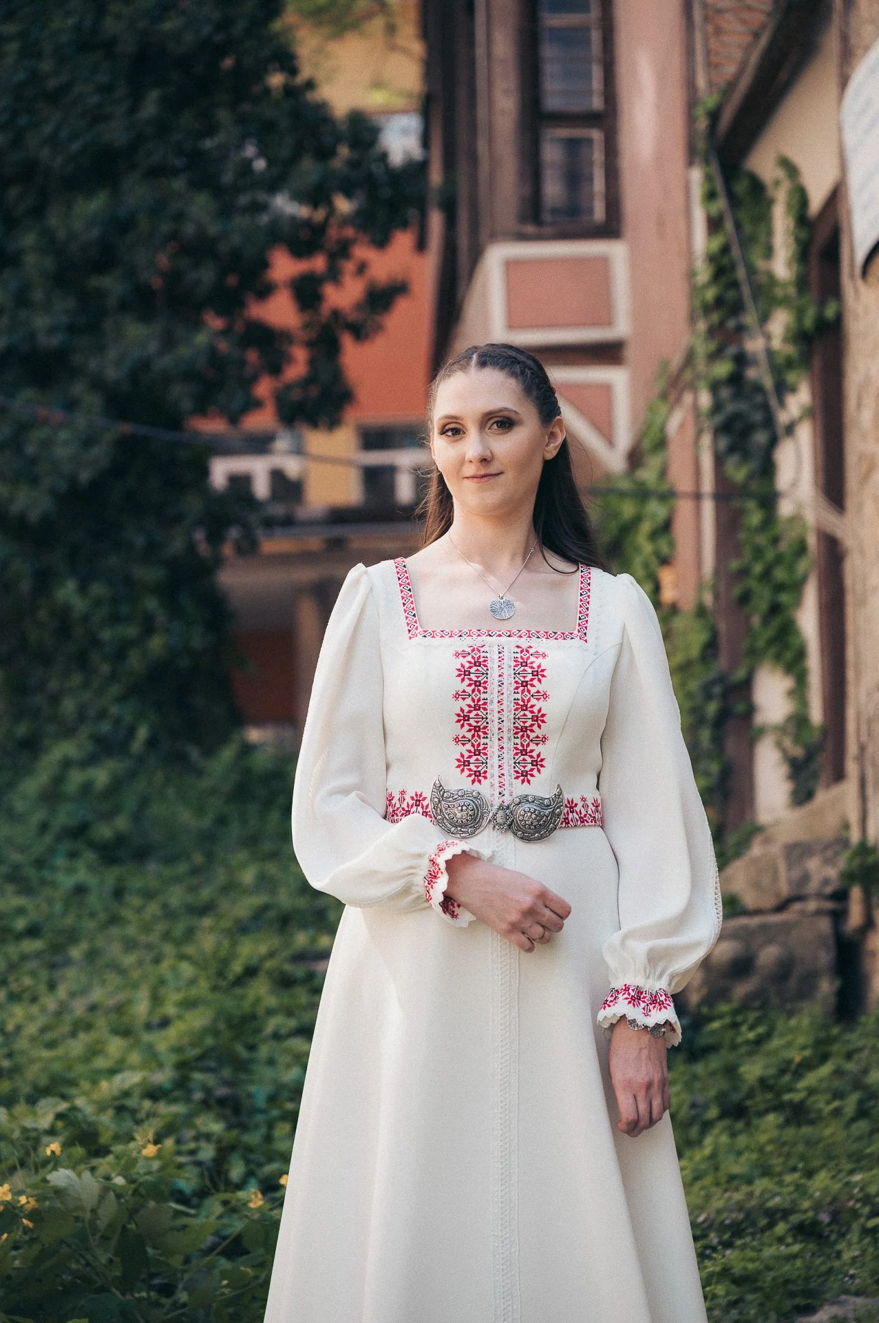 A young woman in traditional Eastern European embroidered dress standing outdoors in front of greenery and a building.