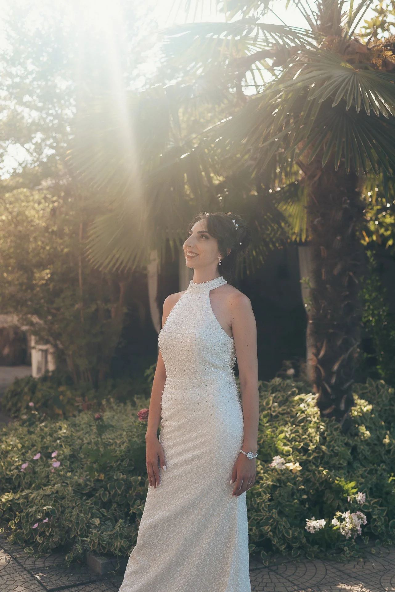Young woman in white wedding dress standing outdoors, smiling, surrounded by green plants and palm trees, sunlight streaming down.