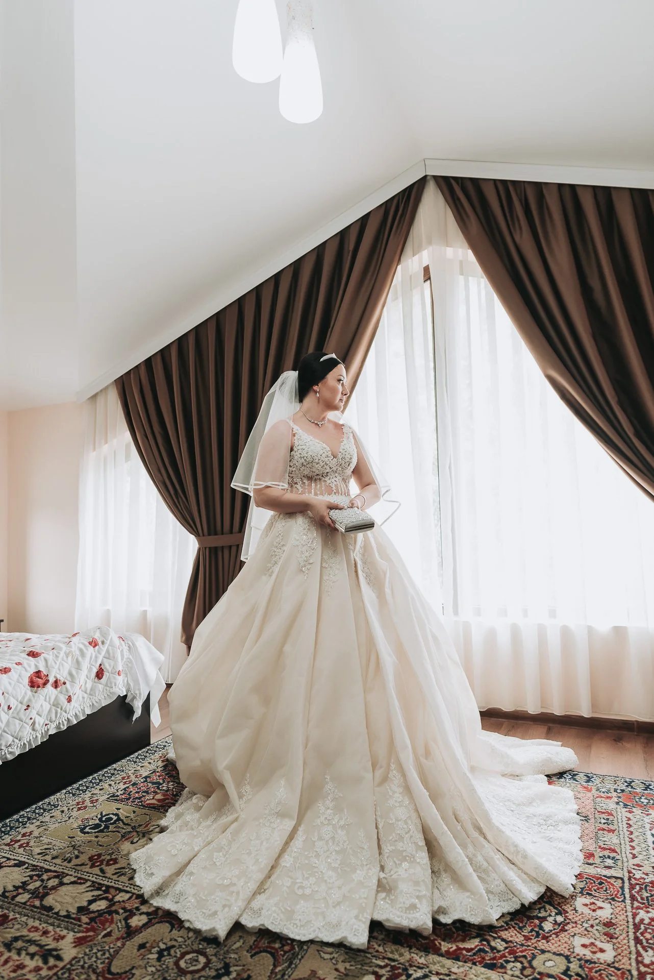 Bride in a wedding gown standing near large windows with brown curtains, holding a clutch purse, in a room with a bed and patterned rug.